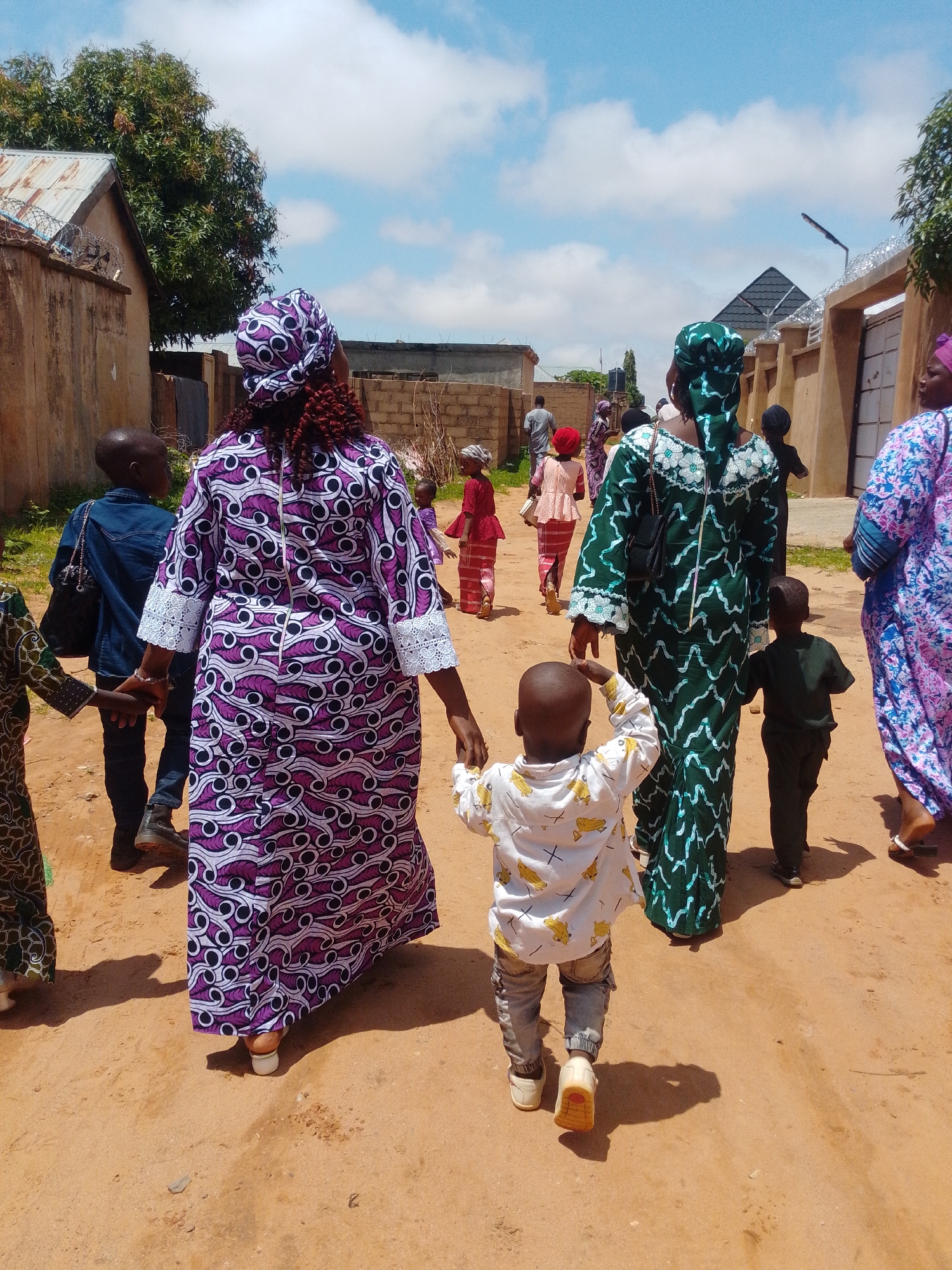 In the city of Tumfure, Nigeria, a group of people walks down a dusty, unpaved street under a bright, partly cloudy sky. The group consists of several women and children, some of whom are walking together.
In the foreground, a young boy in a white shirt with a yellow banana print and grey pants walks away from the camera, holding hands with a woman. The woman is wearing a traditional purple and white patterned dress and a matching headscarf. Behind them, a boy in a denim jacket walks to the left, also holding hands with the woman.
Further back, a woman in a green patterned dress walks with a young boy beside her. Several other individuals, including women in colorful dresses and headscarves, and more children, are visible walking further down the street. The environment appears to be a residential area, with buildings and fences lining the street. A street lamp is visible on the right side of the image. The ground is covered in dry, reddish-brown earth.