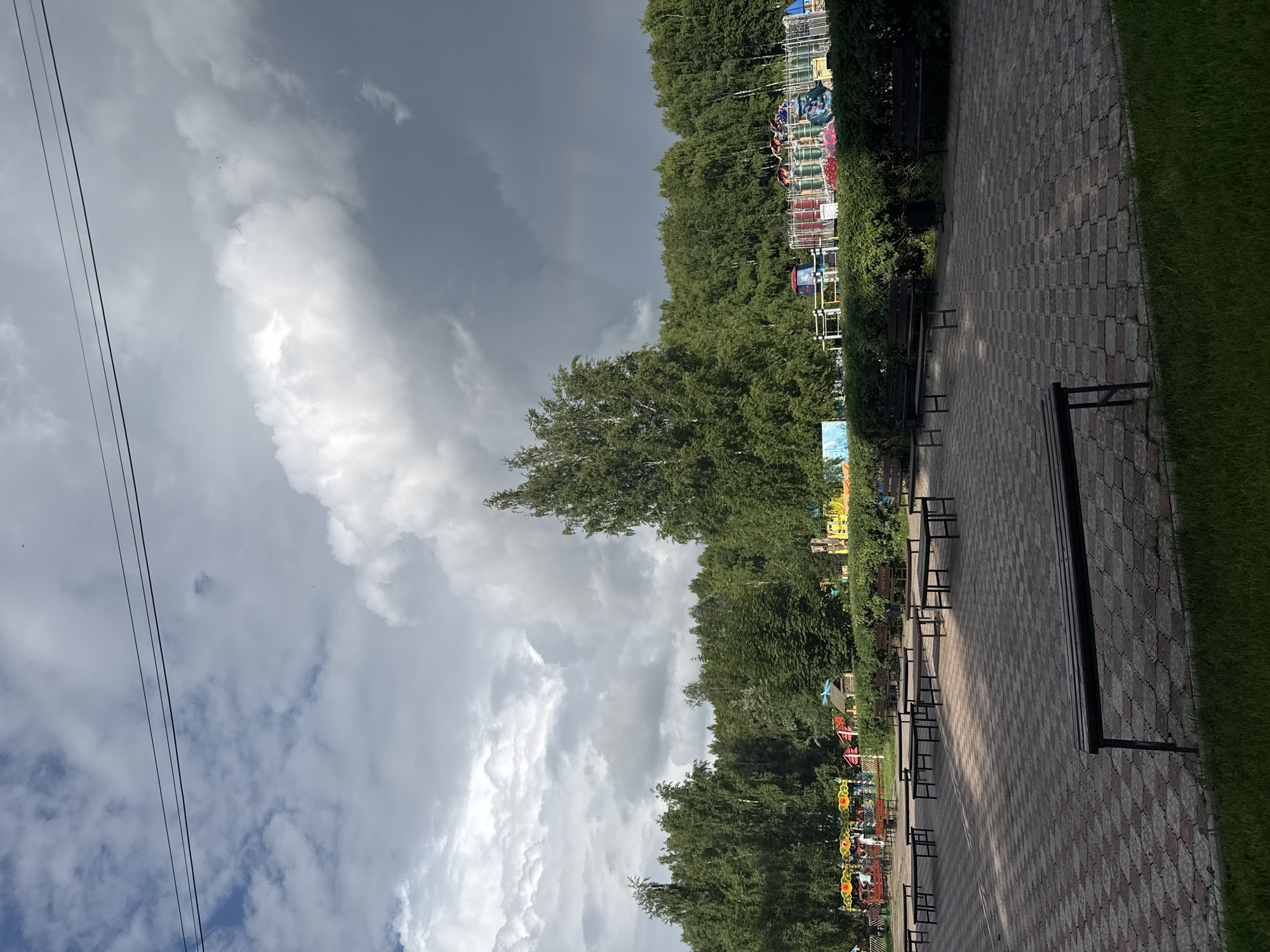 An outdoor amusement park scene is captured on a cloudy day, with a hint of a rainbow visible in the upper right quadrant. The foreground features a paved area with several dark-colored benches and picnic tables scattered across it. Beyond this paved area, a line of green bushes and hedges separates the park grounds from a row of amusement park rides, characterized by colorful structures and netting. A dense line of tall green trees forms the background, extending across the width of the image. The sky is dominated by dramatic, dark gray clouds, with patches of lighter clouds and blue sky peeking through. A few dark power lines are visible stretched across the top of the frame. There are no people clearly depicted in the image. The overall impression is that of a potentially stormy but vibrant amusement park environment.
