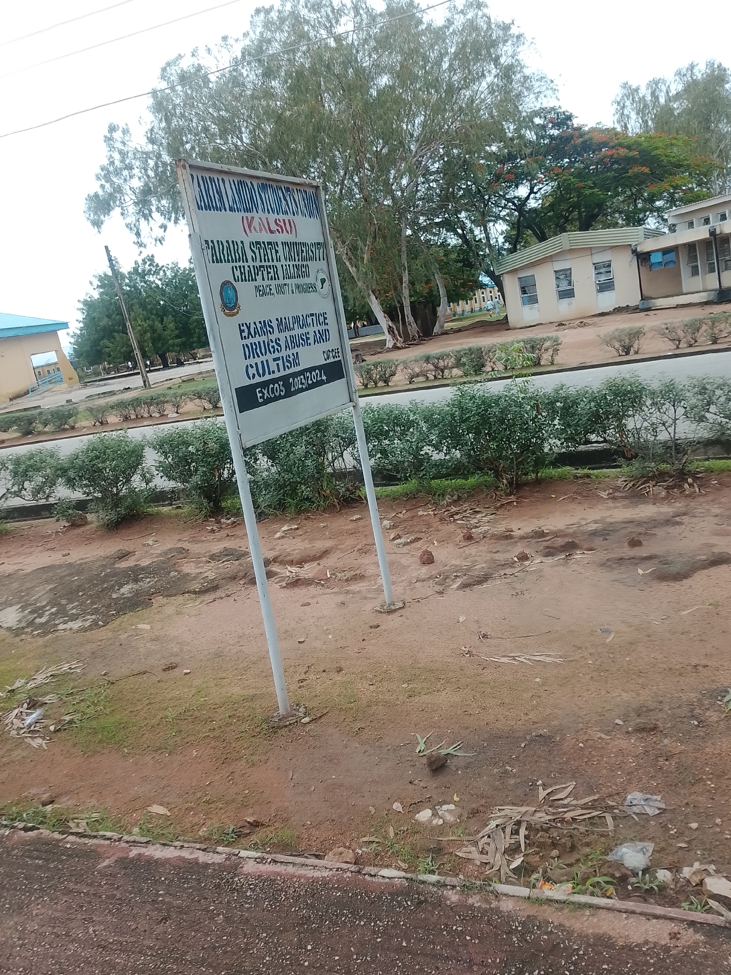 The image shows a signpost in front of some buildings, likely on a university campus. The signpost is white with text in blue and black. The text on the sign reads: "TARABA STATE UNIVERSITY CHAPTER JALINGO PEACE, UNITY & PROGRESS" and then lists "EXAMS MALPRACTICE DRUGS ABUSE AND CULTISM EXCOS 2023/2024". This indicates the location is Taraba State University in Jalingo, Nigeria.
The signpost is supported by two metal poles and is weathered. In the background, there are several buildings, one of which is a light-colored building with windows and a green trim. There are also trees with green foliage and some flowering trees with red blossoms. The ground in front of the signpost and buildings is dirt with some patches of grass and scattered leaves and debris. A paved area or road is visible behind some low-lying green bushes in the middle ground. The sky is overcast. There are no people visible in the image.