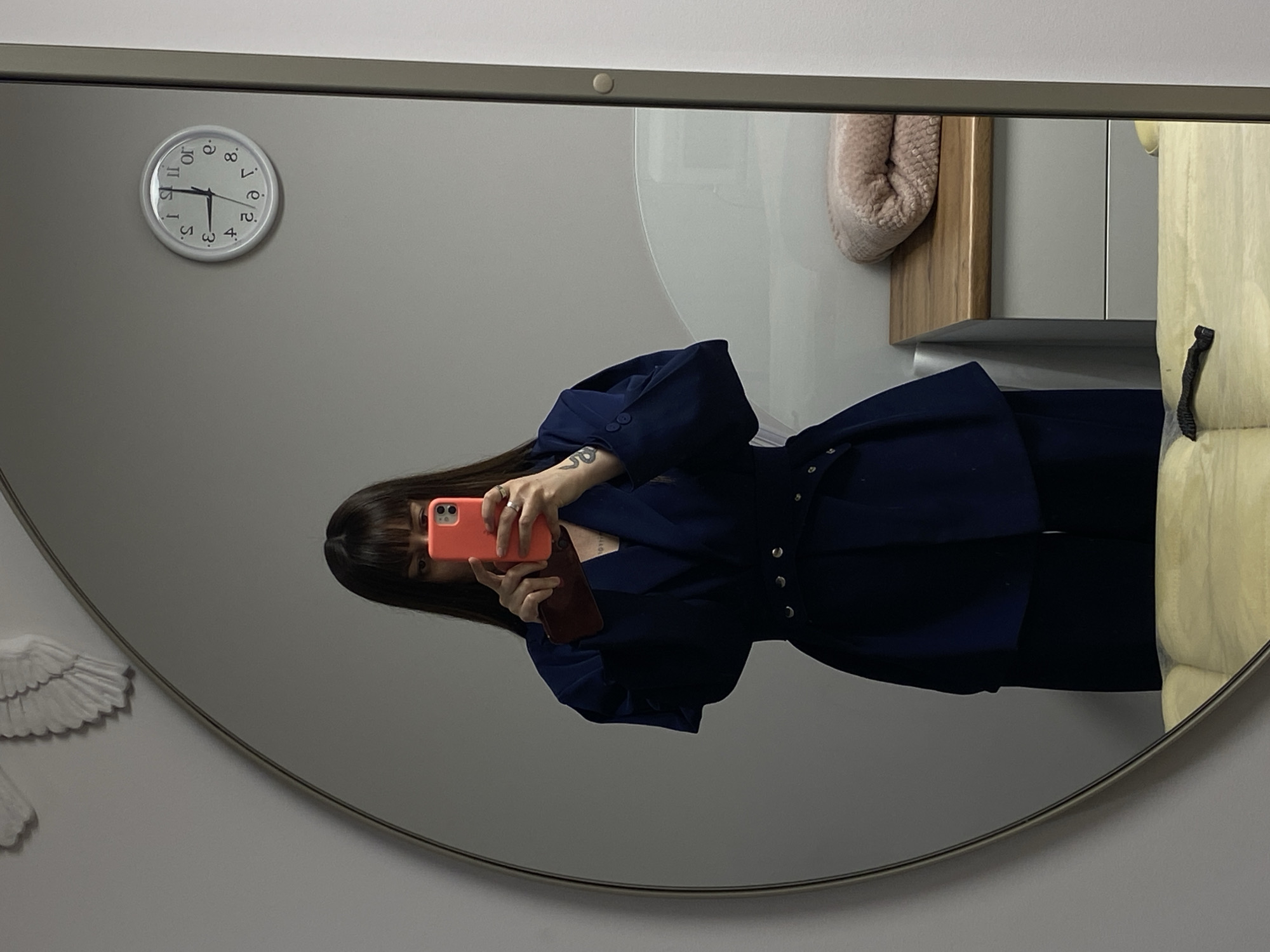 An indoor mirror selfie captures a person in Votkinsk, Russia. The image is taken in a round mirror with a thin metallic frame, reflecting the individual and parts of the room. The person, with long dark hair and bangs, is holding a bright orange smartphone with their right hand to take the picture, partially obscuring their face. A silver ring is visible on their right hand. In their left hand, they hold a dark brown wallet or cardholder. The person is wearing a dark blue garment, possibly a jacket or dress, with distinctive puffy sleeves and a line of buttons down the front. A tattoo, possibly a snake design, is visible on their left arm.
On the wall above the mirror, a white circular clock is reflected, showing the time as approximately 9:00, with the minute hand pointing to 12 and the hour hand pointing to 9. The clock's numbers are visible but appear mirrored. To the left of the mirror, a white decorative object resembling an angel's wing is affixed to the wall. In the background reflection within the mirror, a light-colored, possibly pink, textured blanket or towel is seen draped over a wooden shelf or cabinet. To the right, a section of a light-colored, perhaps quilted, piece of furniture is visible. The walls appear to be a neutral light gray or off-white color.