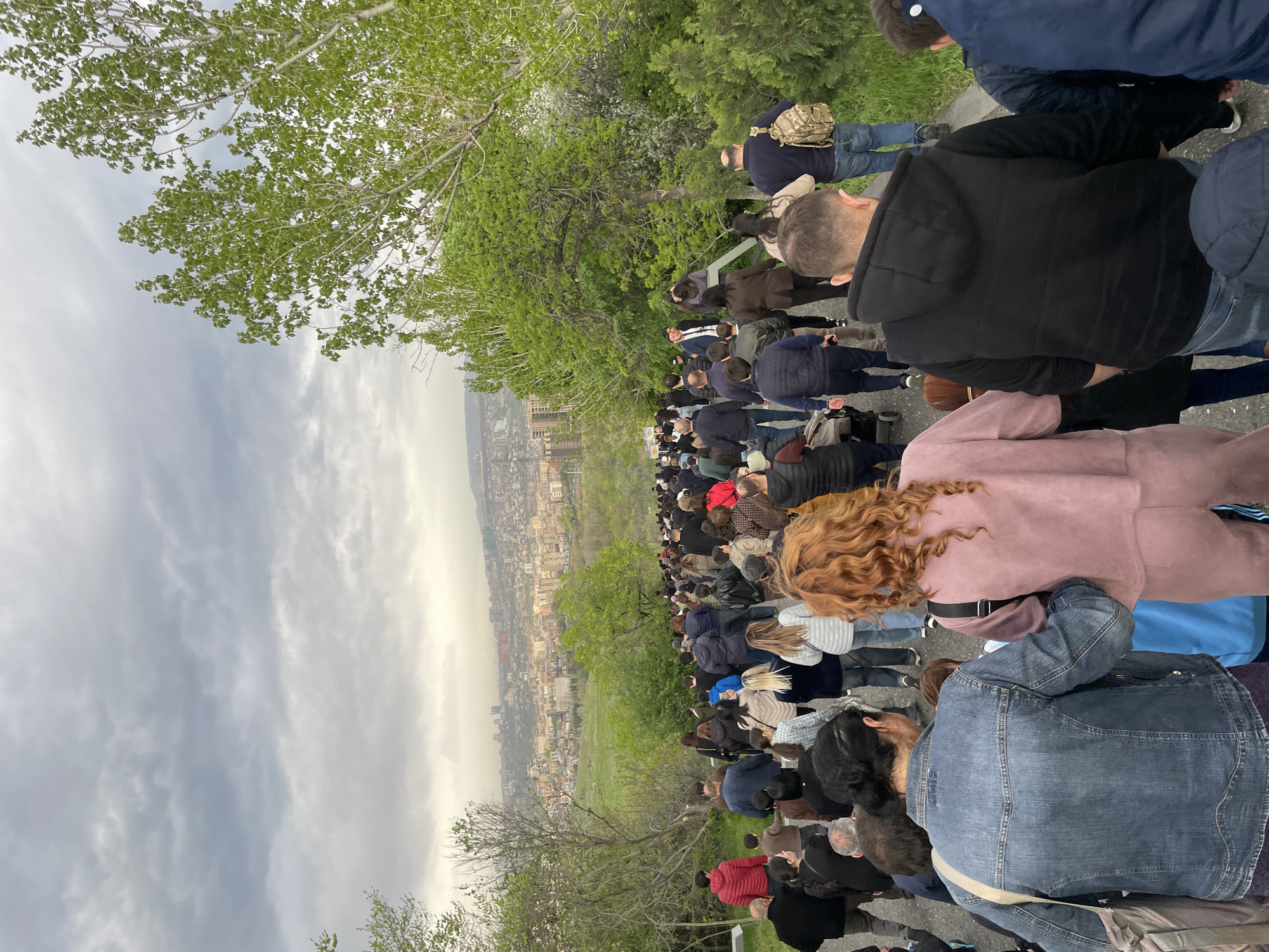 The image captures a large outdoor gathering of people, viewed from a slightly elevated perspective. The photograph is oriented vertically, with the left side of the frame serving as the top. A dense crowd, predominantly composed of adults, fills the midground and foreground. Many individuals are seen from behind, facing away from the camera and towards a distant cityscape. In the immediate foreground, a woman with long, curly auburn hair and a pink jacket, and another person in a denim jacket, are prominent. The crowd extends far into the midground, with people wearing various casual clothes, including jackets and hoodies.
The setting appears to be a hillside or an elevated park overlooking an urban area. Lush green trees with broad leaves are visible on the left side of the frame, and more scattered greenery is seen between the crowd and the city. The background reveals a sprawling cityscape with numerous buildings, stretching across the horizon under an overcast or partly cloudy sky. The lighting suggests daytime, possibly late afternoon or early evening, with a diffused light that creates a somewhat muted atmosphere. The location, given the context, is Yerevan, Armenia.
The activity appears to be a public event or assembly, as indicated by the large number of people gathered and looking in a common direction. There is no clear indication of specific emotions, but the collective focus suggests a shared purpose or observation. No legible text is visible in the image.