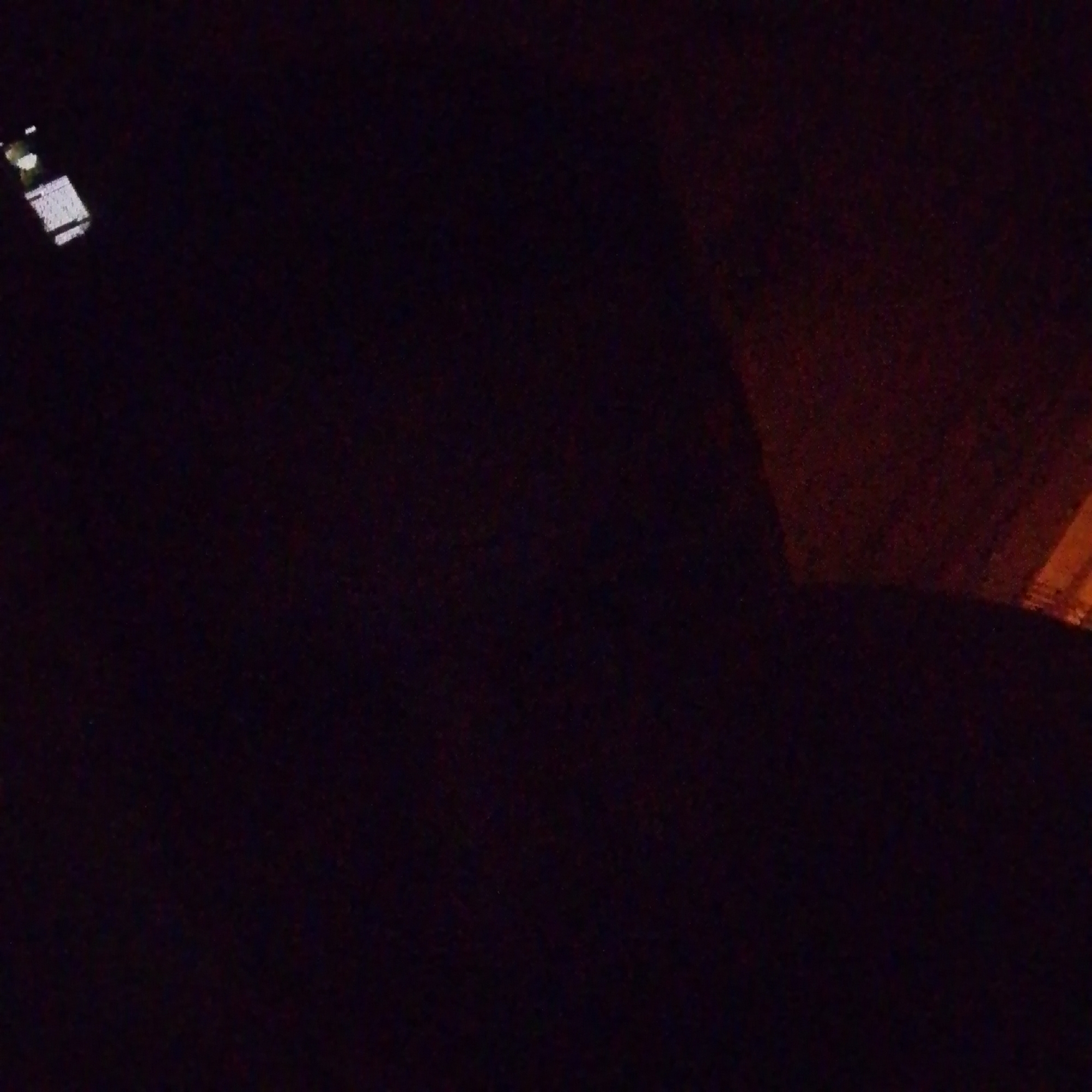 The image depicts a dimly lit scene, possibly taken at night or in a poorly illuminated indoor space. In the upper left portion of the image, a dark rectangular object with a lighter rectangular screen is visible. This object appears to be a mobile phone or a similar electronic device. The screen shows several bright horizontal lines, suggestive of a keyboard or text display. The rest of the image is predominantly dark, with a faint reddish-brown glow emanating from the upper right side. This glow suggests an artificial light source, potentially a streetlamp or a light from a building, casting a warm, diffused light on a textured surface. The overall impression is one of low visibility and a potentially urban environment, though the specific location in Malye Vyazemy, Russia, cannot be confirmed from the visual information alone. There are no people or discernible actions visible in the image.