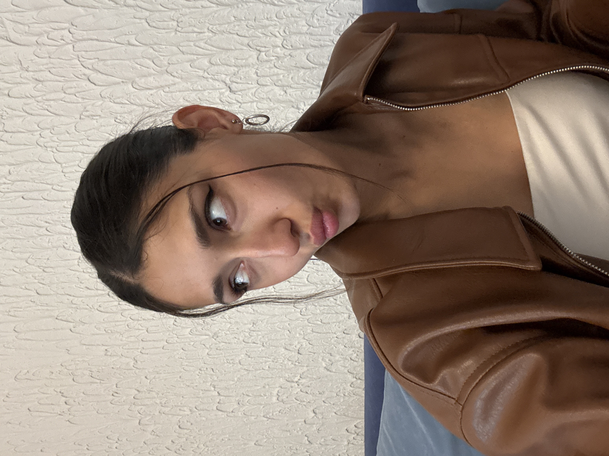 A woman with dark hair pulled back is wearing a brown leather jacket over a white top. She is facing the camera and appears to be making a pouty face. She is wearing a silver earring in her left ear. The background is a textured off-white wall. A blue object is partly visible behind the jacket.
The location in Zankovići, Montenegro is not discernible from the image.
