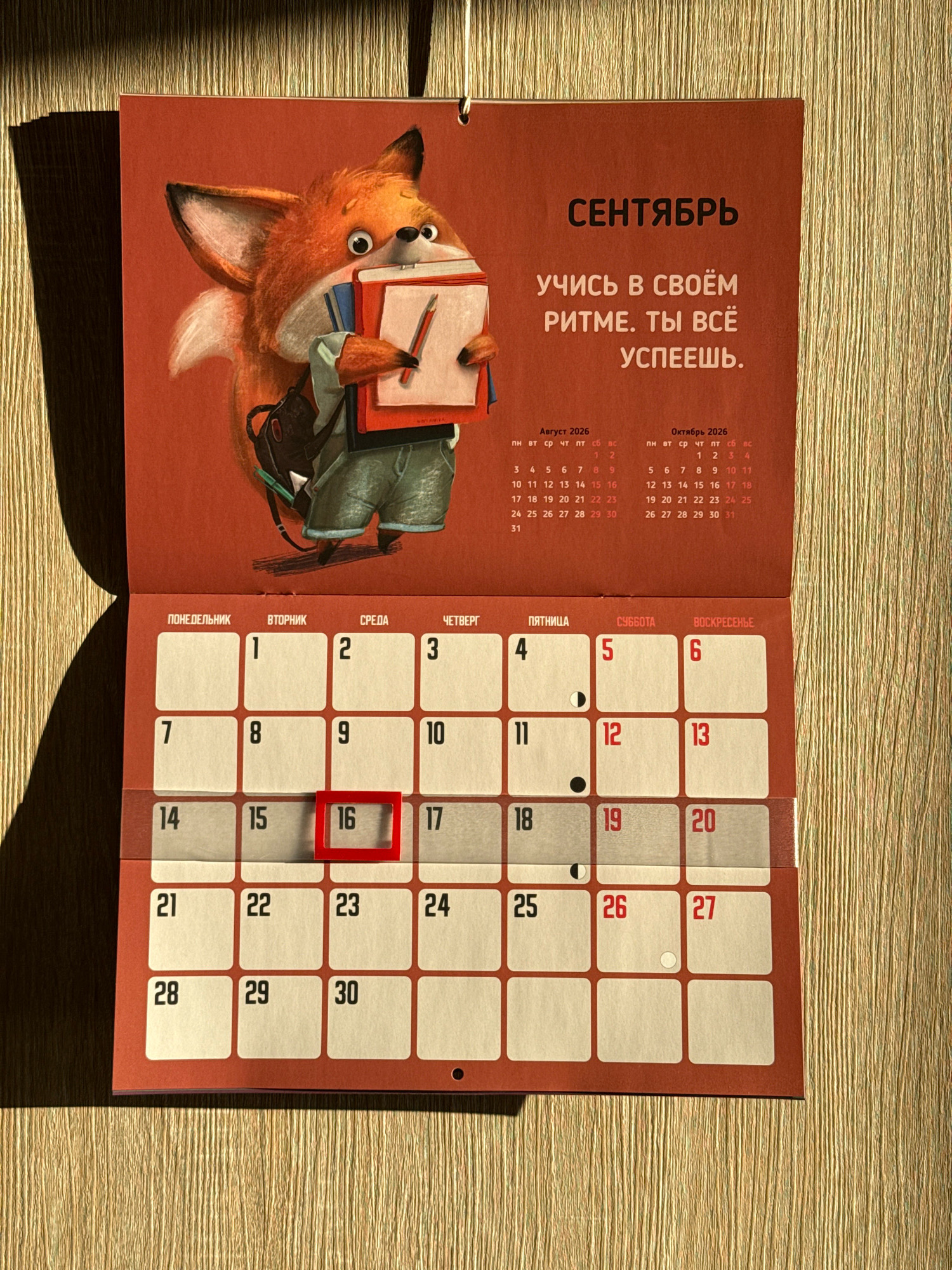 The image is a close-up shot of a wall calendar featuring a cartoon fox character. The calendar is predominantly red and features the Russian text "СЕНТЯБРЬ" (September) and an inspirational quote. Below the illustration, there is a grid calendar for September. The date "16" is highlighted with a red border, indicating it is the current or an important day. The calendar is attached to a textured wall, with a shadow cast across the left side of the image. The background appears to be a wooden surface with vertical lines. There are no people or specific landmarks visible that would allow for a precise location within Lyubertsy, Russia.