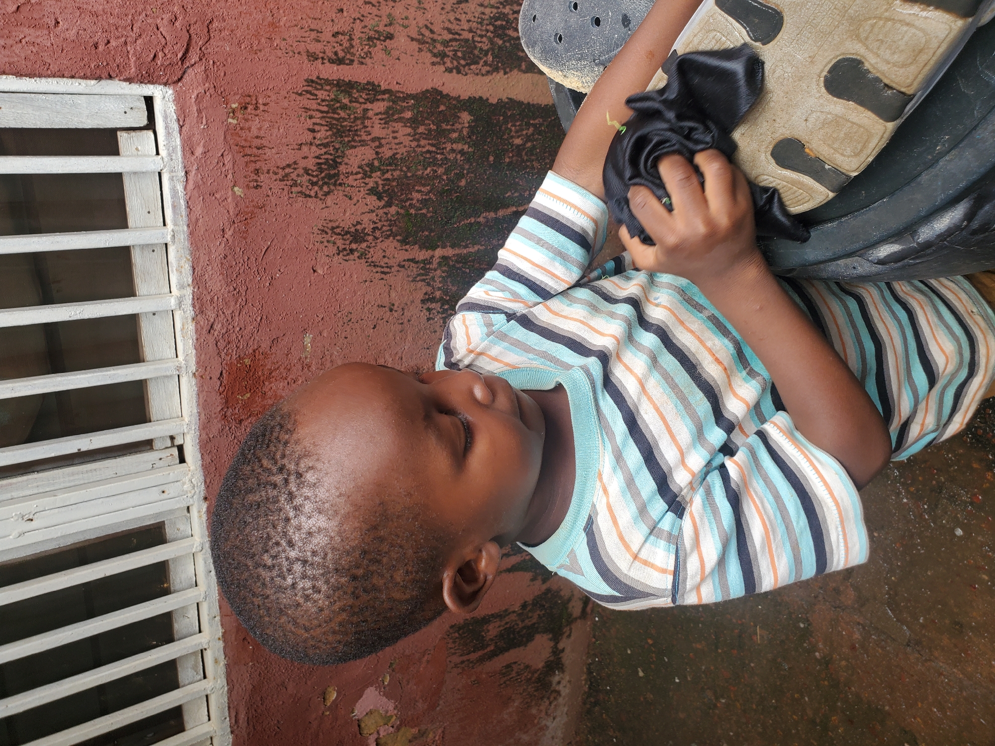 A young child, likely a boy due to his short hair and clothing, is lying on a reddish-brown textured surface, possibly concrete or stucco. He is wearing a striped t-shirt with horizontal bands of blue, orange, gray, and black. His eyes are closed, suggesting he is resting or sleeping.
His right hand is holding a black piece of fabric, which is bunched up. His hand and the fabric are resting on the sole of a large, dark-colored shoe or boot, which has a textured grip pattern. The shoe appears to be worn.
To the left of the child, there is a white window grill with horizontal slats. The wall behind the grill is also reddish-brown and appears weathered, with some dark patches, possibly moss or dirt. The ground the child is lying on also shows some dark, mottled patches, indicative of dampness or organic growth.
The scene appears to be outdoors, possibly in a backyard or patio area. The lighting does not suggest a specific time of day, but the overall impression is of a casual, everyday moment. There is no visible text in the image. The setting could be a residential area in Jos, Nigeria, given the context provided. The child's expression is serene, suggesting a peaceful state.