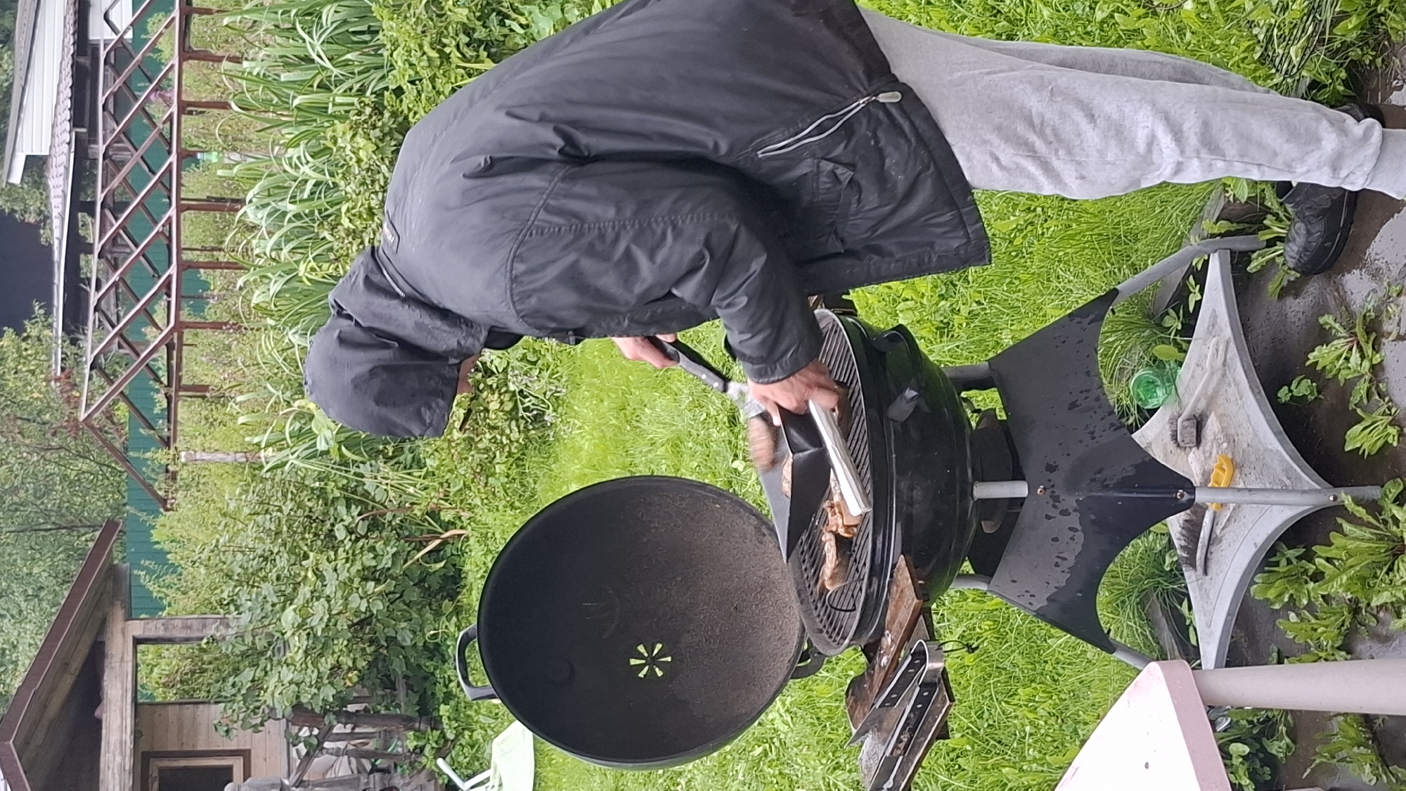 A person wearing a black hooded jacket and gray sweatpants is grilling food on a black charcoal grill in a grassy yard. The grill is open, revealing meat on the grates. The person is holding tongs and a black grilling tool, appearing to turn or move the food. In the background, there is a wooden structure resembling a small shed or outbuilding, a greenhouse with a metal frame, and lush green vegetation. The ground appears wet, suggesting recent rain. The scene is set in a rural or semi-rural outdoor environment.
