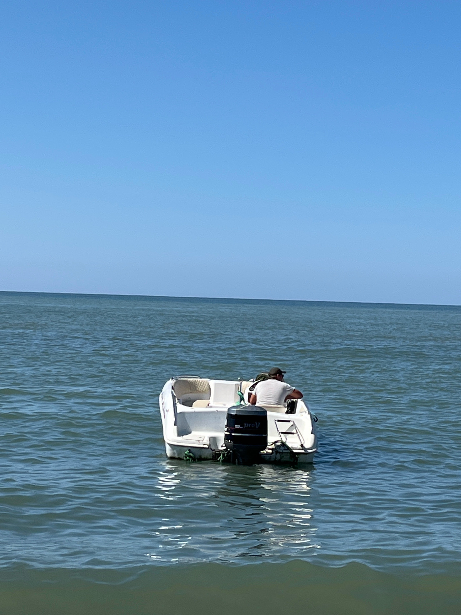 A person is seated in a white motorboat in the sea. The person is wearing a light-colored t-shirt and a dark baseball cap, facing away from the camera and towards the water. The boat has an outboard motor at the stern. The sea is calm with gentle ripples, and the sky is clear and blue. The horizon is visible as a thin line separating the sea from the sky. The image appears to be taken from another boat or from the water. This scene could be on the Black Sea coast near Ureki, Georgia, a location known for its beaches and recreational boating.
