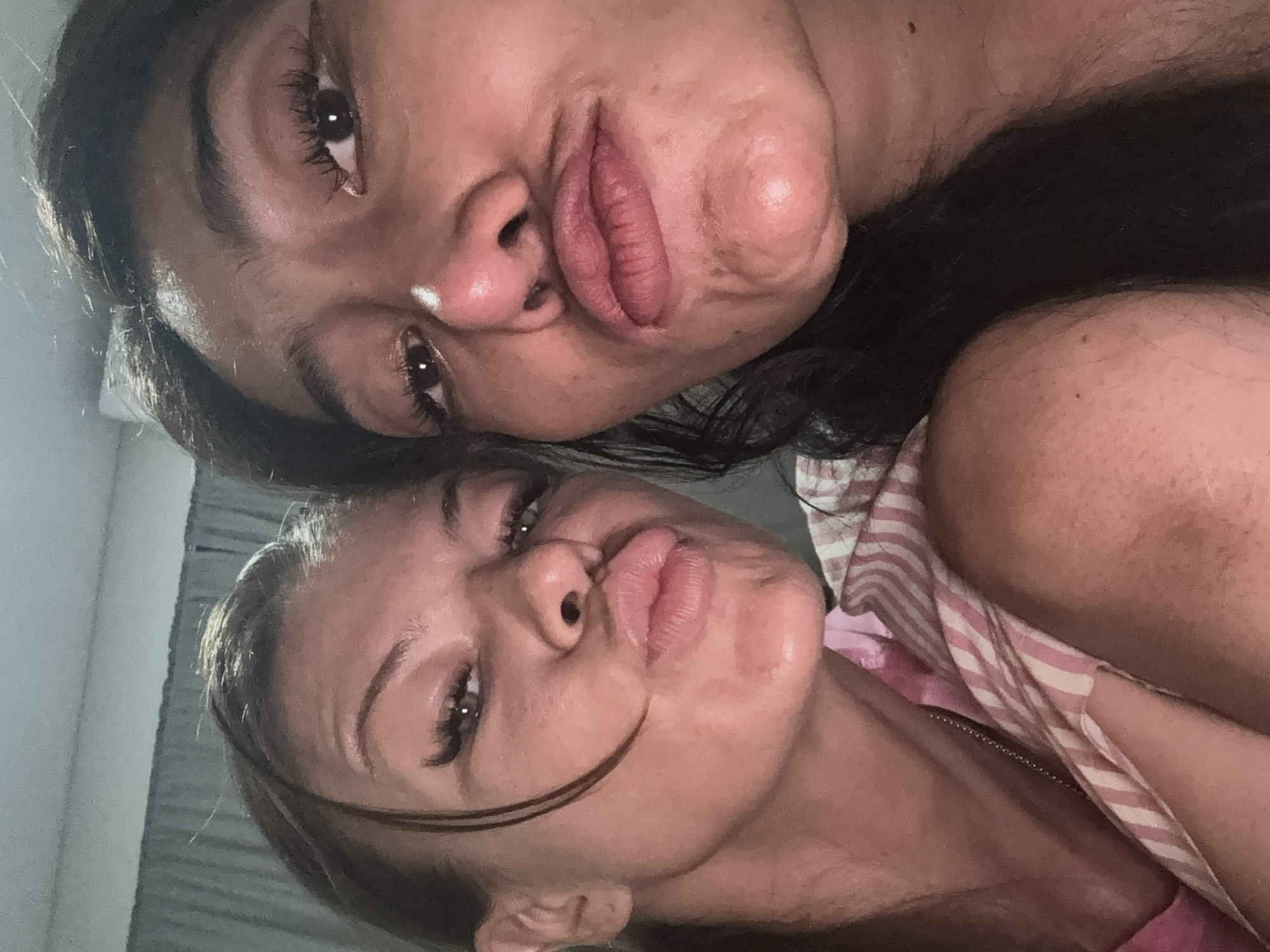 This close-up selfie, captured in Kuta, Indonesia, features two women posing with their faces close together, both looking towards the camera with pouting expressions. The image is oriented sideways.
In the foreground, one woman has a lighter skin tone and light brown hair, with a few strands falling across her face. Her lips are full and prominently pursed, and her eyes feature long, dark eyelashes. She appears to be wearing a pink and white striped top, partially visible on the right side of the frame.
Behind and slightly above her, the second woman has a darker complexion and dark hair. Her lips are also full and intentionally pursed, mirroring the pose of the first woman. She has notable long eyelashes, and a small white highlight or possibly a blemish is visible on her nose.
The lighting is dim, suggesting an indoor setting or evening conditions, and the background is largely dark and out of focus, providing minimal environmental context beyond the subjects. Both women convey a playful or stylized mood through their expressions. No text is visible in the image.