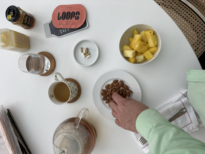 An overhead shot captures a white table surface in the City of London, United Kingdom, presenting a collection of food and beverage items. A light-skinned hand, clad in a light green long-sleeved shirt, reaches to pick an almond from a white plate of whole almonds on the right. Nearby, a white napkin with red embroidered trim holds a metal fork. A white bowl contains diced pineapple chunks, and a small white dish holds several multi-colored capsules. Beverages include a clear glass teapot with amber liquid, a mug of amber liquid on a cork coaster, and a glass of water, also on a cork coaster. Complementing these are a small jar of Manuka honey, a clear bottle of light-colored liquid, and a reddish-pink product packet labeled 'LOOPS.' A rattan chair back is partially visible in the upper right. Stacked papers or magazines are partially visible on the far left.