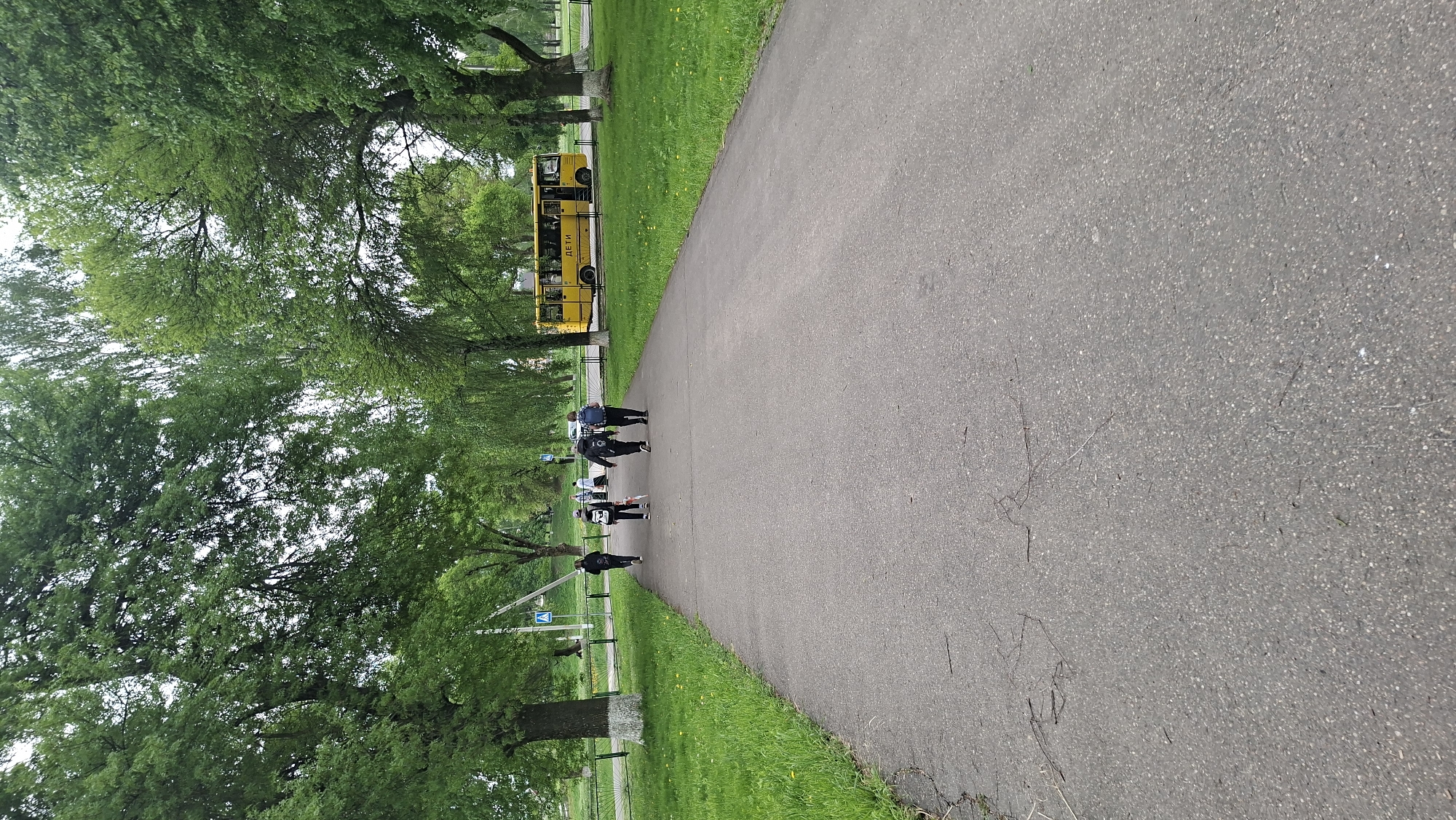 A long, asphalt path winds through a green park. On the left side of the path, there are mature trees and grassy areas. A yellow bus is parked in the background. Several people are walking along the path in the middle distance. The scene appears to be in Забалоцкі сельскі Савет, Belarus.
