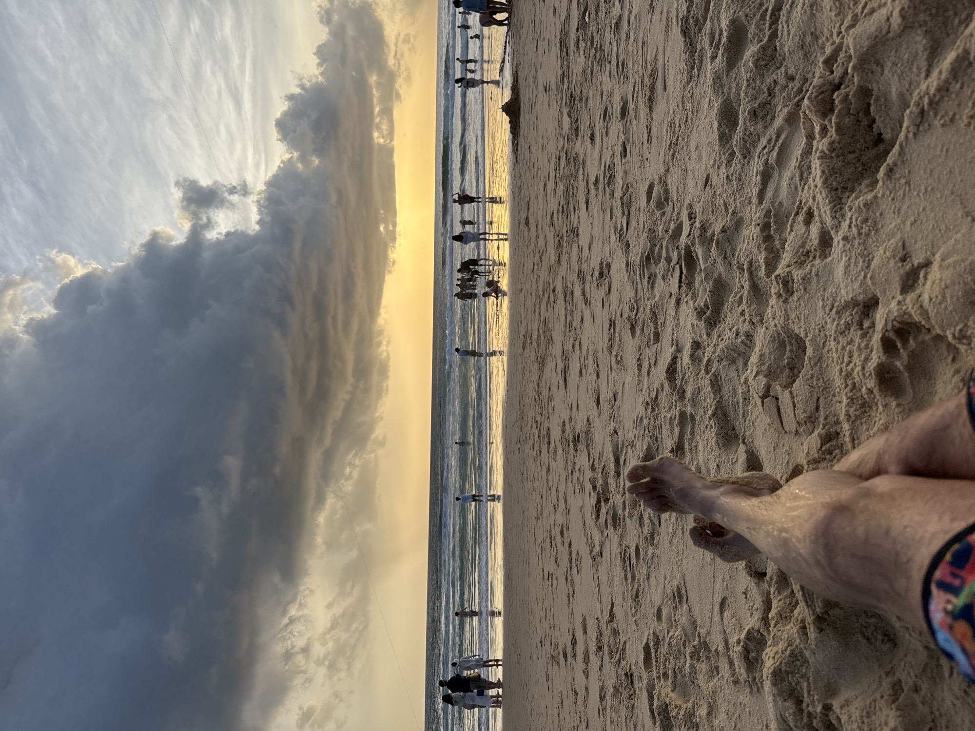 This image captures a sunset scene at Karon Beach in Karon, Thailand. The foreground is dominated by a close-up view of a person's legs and feet resting on the sandy beach, with colorful swim trunks visible. The sand is textured, showing footprints and ripples. In the midground, the ocean meets the shore, with gentle waves rolling in. Several people are scattered along the water's edge and in the shallow sea, engaged in various activities like standing, walking, and playing. Some are silhouetted against the setting sun. The background features a dramatic sky filled with dark, heavy clouds interspersed with patches of golden light, indicating the sun is low on the horizon. The overall impression is one of a relaxed evening at a popular tourist beach.