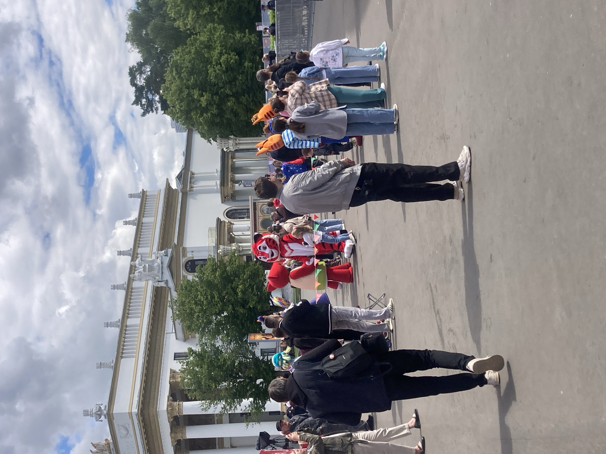 The image shows a crowd of people standing in a line outside a white building with statues on top, potentially in Moscow, Russia. There is a mascot character in the middle of the line. The sky is cloudy.