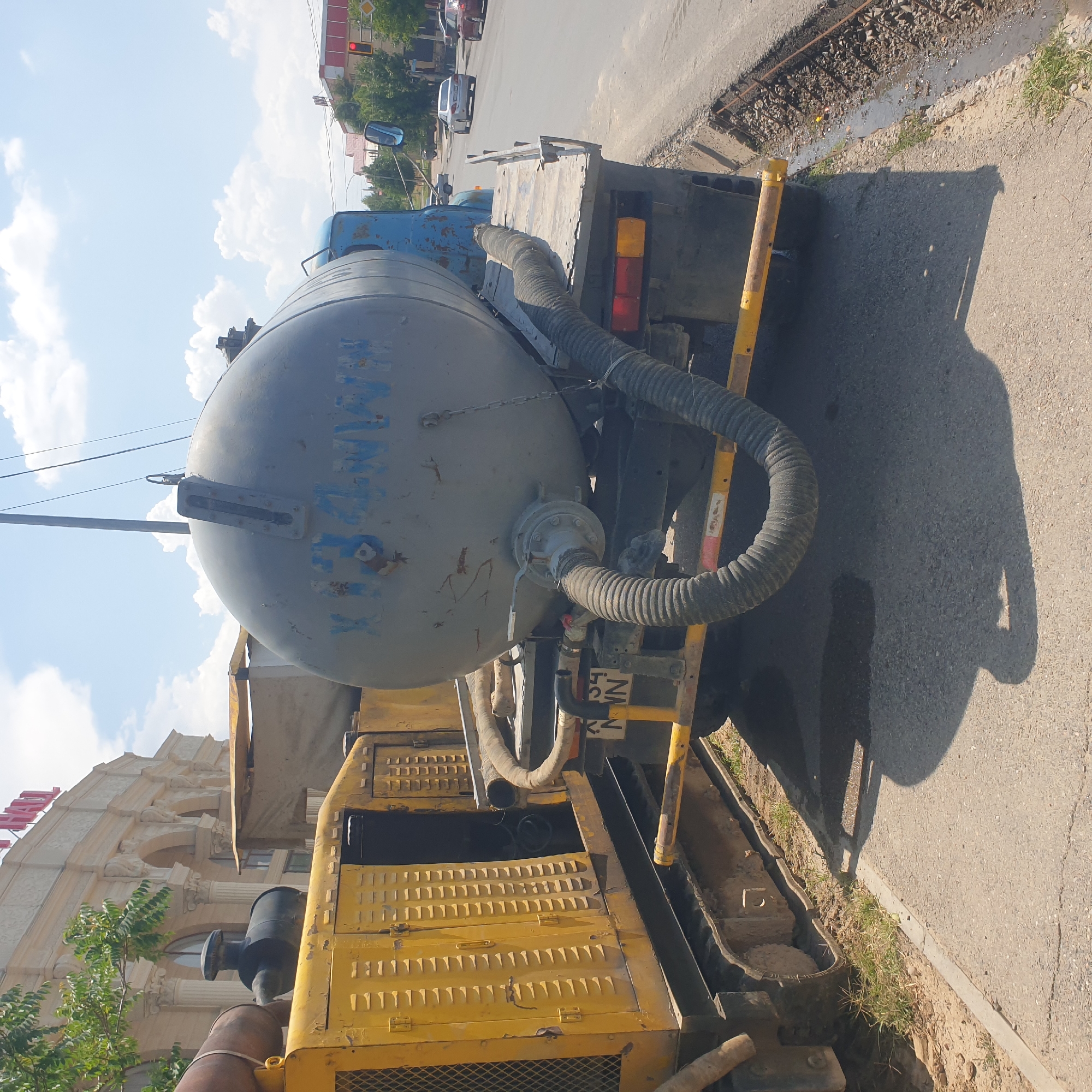 A yellow construction vehicle and a blue truck with a large grey tank are parked on the side of a street in Shymkent, Kazakhstan. The truck has a license plate that reads "K134 NN". The vehicles appear to be part of a road construction or maintenance project, as there is disturbed ground and gravel along the roadside. In the background, there is a large building with arched windows and columns, and other vehicles are visible on the street. The sky is blue with scattered clouds.