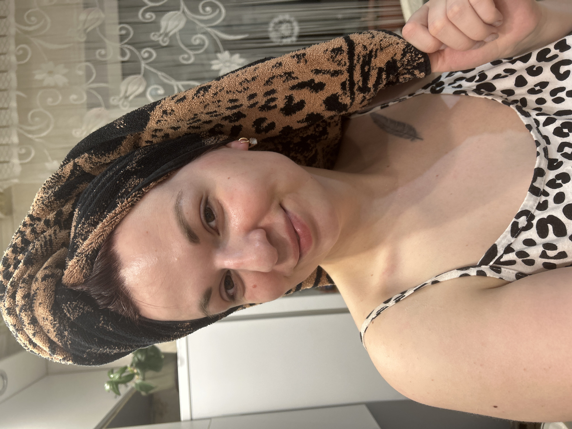 A woman poses for a selfie, a patterned towel wrapped around her head, wearing a black and white patterned top. A feather tattoo is visible on her chest. The background includes a floral patterned curtain, a white refrigerator, and a potted plant. The location is unknown.