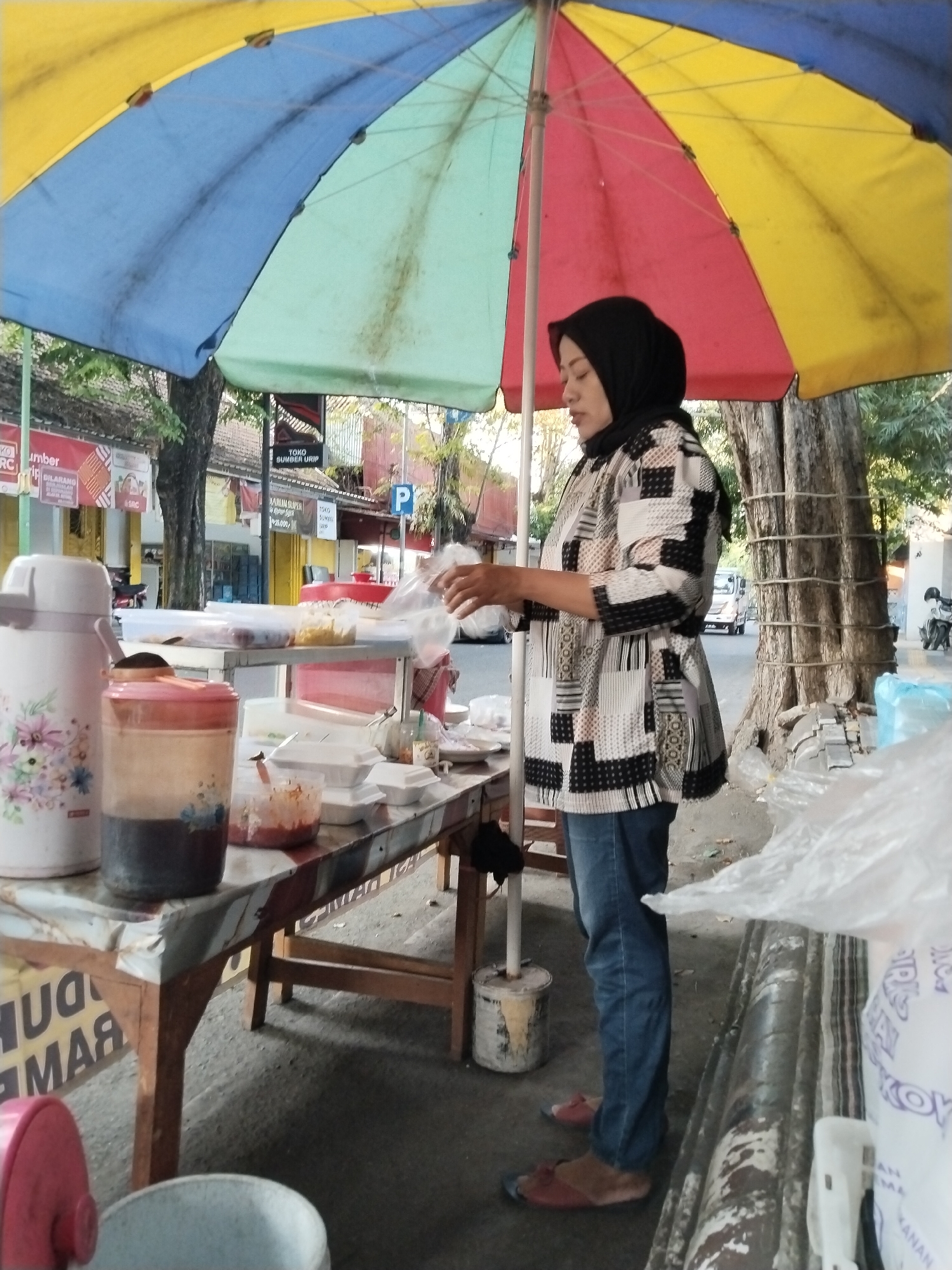 A woman wearing a black hijab and a black and white patterned shirt is standing at a food stall under a colorful umbrella. She is holding a clear plastic bag, appearing to be packing food. The stall has a wooden table covered with a patterned plastic cloth. On the table, there are several white food containers, a large thermos with a floral design, and two plastic jars, one containing dark liquid and the other with a reddish substance. In the background, buildings and trees are visible, suggesting an outdoor urban setting. A street with a passing vehicle can be seen behind the stall. The overall scene depicts a typical street food vendor operation in Kecamatan Pati, Indonesia.