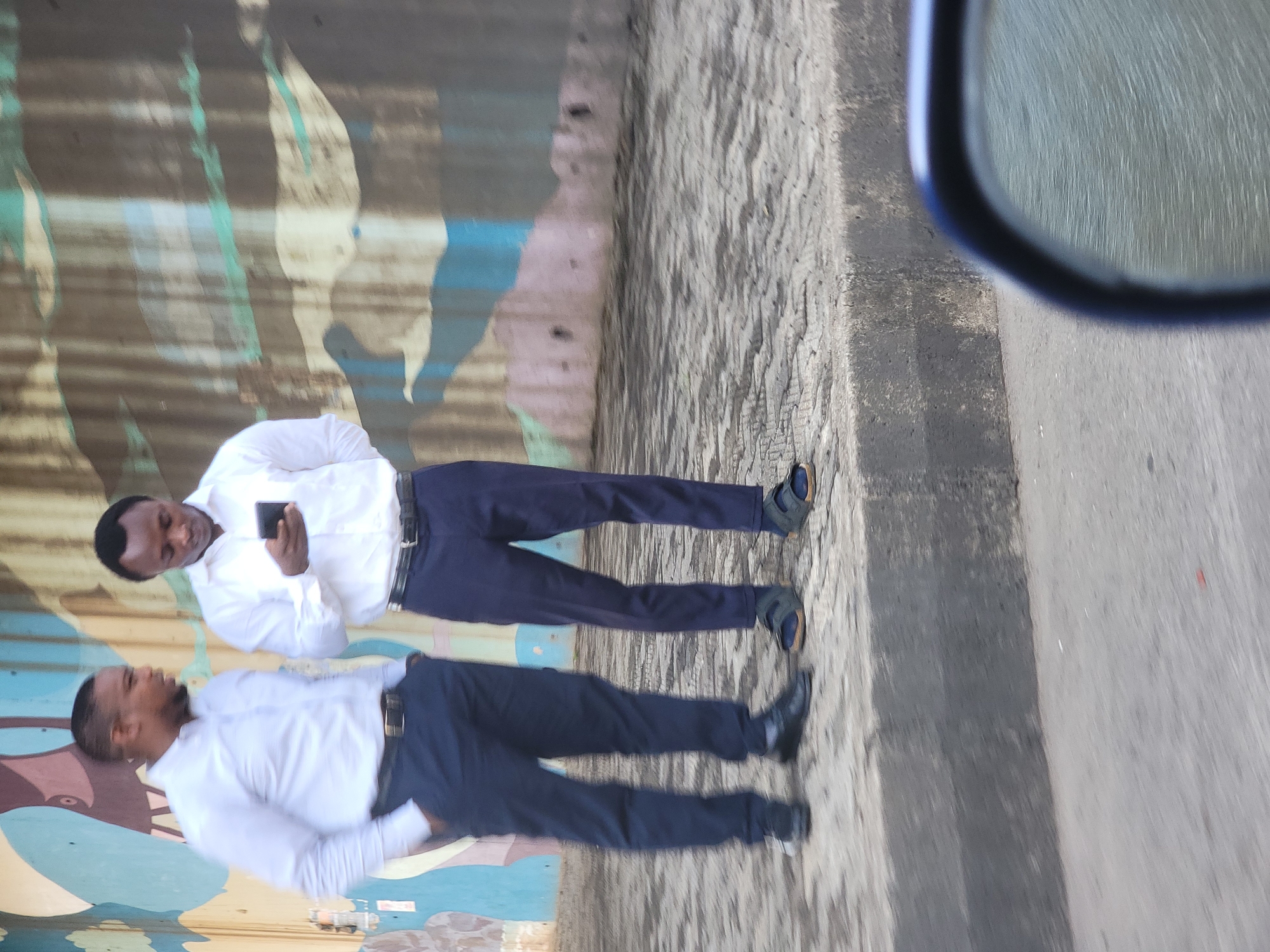 Two men stand side-by-side on a sidewalk in Jos, Nigeria. The man on the right is holding a mobile phone and looking at it. Both men are wearing white shirts and dark trousers. The background includes a corrugated metal wall with painted colors and a gray concrete wall with a road. A vehicle mirror is visible in the top right.