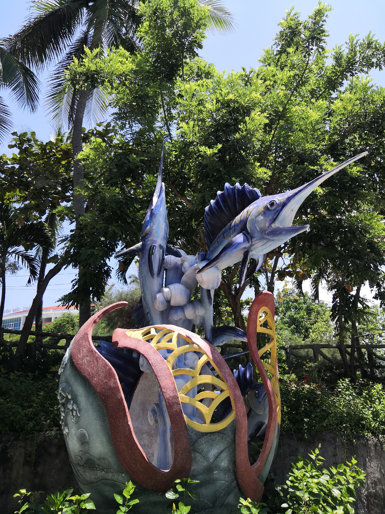 The image displays a vibrant, multi-component sculpture situated outdoors in Bac Nha Trang Ward, Vietnam. The central feature is a large, dynamic statue of a blue marlin or sailfish, predominantly colored in shades of blue, white, and grey, depicted mid-leap with its mouth open and a prominent spiked snout. A second, similar fish sculpture is partially visible behind and below the main fish. These fish are integrated into a substantial, multi-layered base structure. This base exhibits curvilinear forms in reddish-brown and golden-yellow, possibly representing waves or abstract marine elements, and contains an internal cavity revealing a smaller fish statue within.
The sculpture is set against a backdrop of lush green vegetation, including several tall palm trees with visible fronds on the left and various broadleaf trees filling the upper and mid-ground. A clear, bright blue sky is visible above the foliage. In the far left background, partially obscured by trees, a light-colored building with multiple rectangular windows and a red roof is discernible. Text, partially legible as "TÔN ĐỨC TH," is visible on a sign associated with this building. A rustic-style fence constructed from wooden logs or branches spans horizontally across the mid-ground, behind the sculpture and amidst the greenery. The foreground features additional green ground cover and foliage. The scene is brightly illuminated by natural sunlight; no human figures are present.