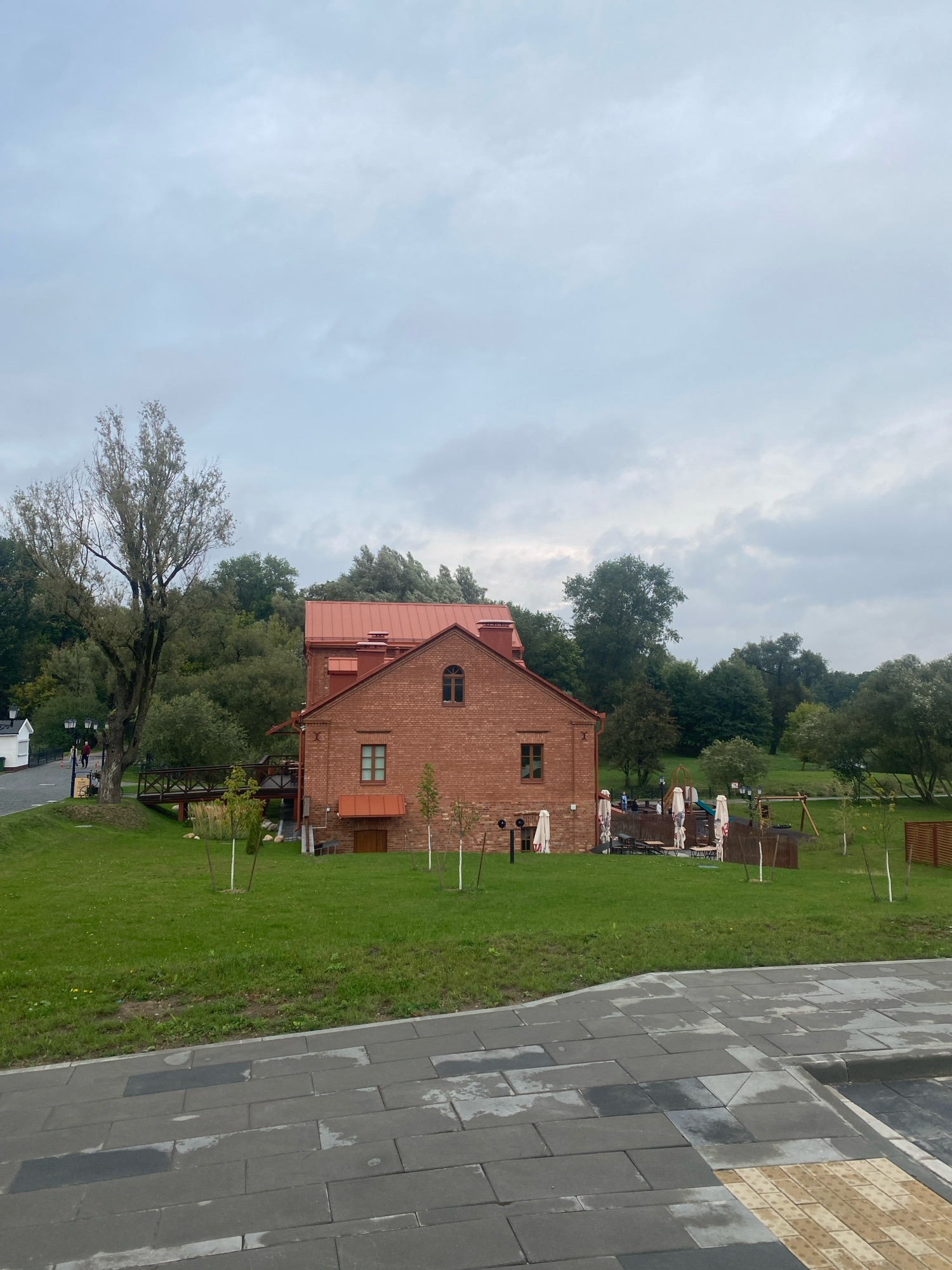 The image shows a brick building with a red roof in an outdoor setting. The building appears to be a historical structure, possibly a mill, given its architecture. In front of the building is a grassy area with several trees and a playground. There are tables and umbrellas set up on the grass, suggesting an outdoor seating area for a restaurant or cafe. A paved walkway is visible in the foreground. The sky is overcast with clouds. The location is identified as the "Belarusian State Museum of Folk Architecture and Rural Life" (also known as Dudutki) in the Minsk region of Belarus.