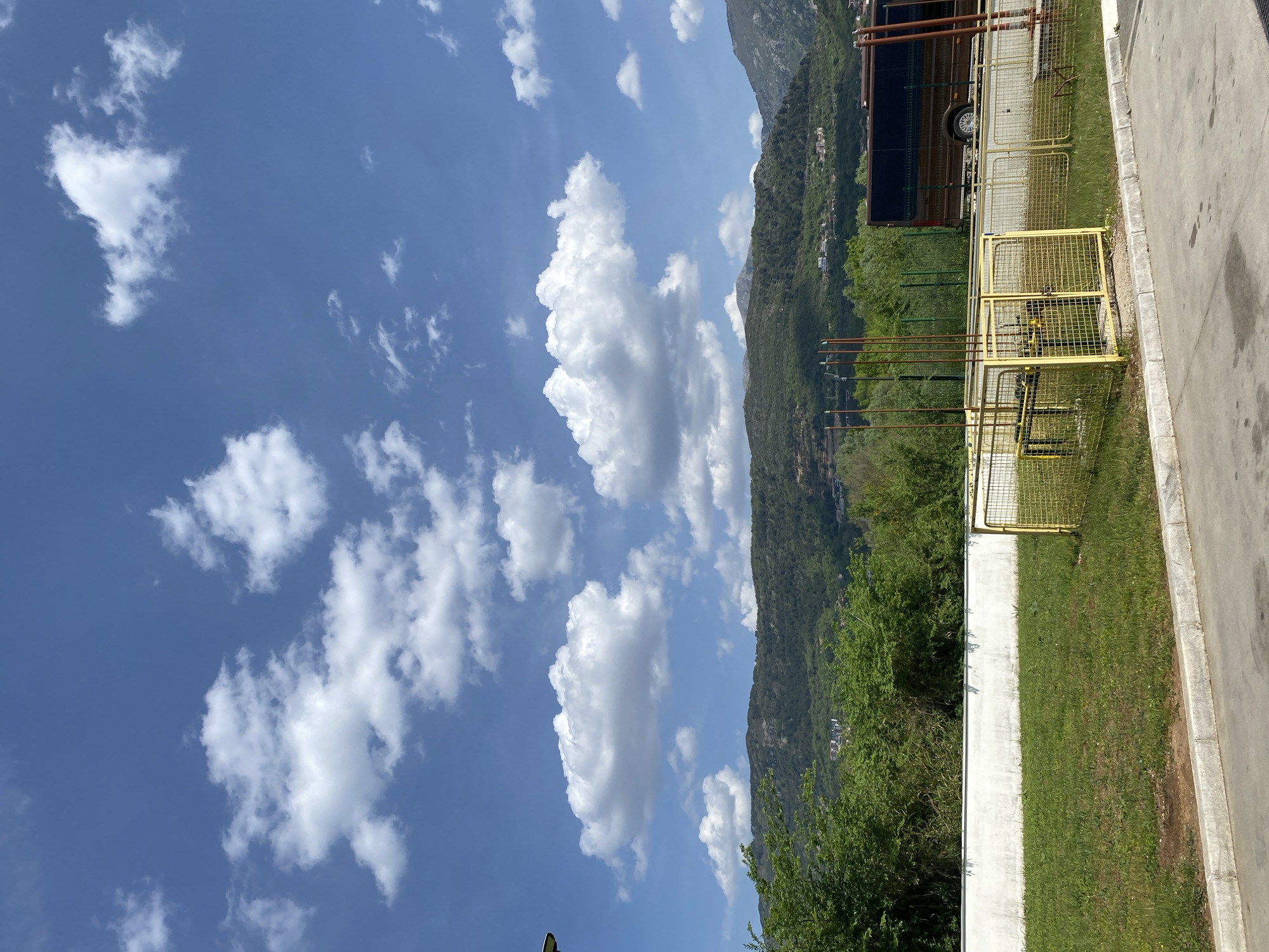 This outdoor image captures a picturesque scene under a bright, sunny sky with scattered white cumulus clouds. The dominant feature in the background is a range of lush, green mountains, heavily forested with trees. Faintly visible on the distant mountain slopes are small buildings, indicating some human settlement.
In the midground, a strip of vibrant green grass runs horizontally, bordered by a white concrete wall or barrier. Behind this barrier and grass, a prominent yellow metal mesh fence or barrier encloses an area. Within this fenced-off section, several tall, brown, pipe-like structures are visible, suggesting an industrial or utility complex, possibly related to gas or water infrastructure. A large, dark brown truck or trailer is partially visible behind the yellow fence.
The foreground on the right features a paved area, likely a road or parking lot, bordered by a concrete curb. No human figures or discernible activity are present in the image. The time of day appears to be bright daylight, given the clear blue sky and strong illumination. There is no visible text. The scene offers a blend of natural beauty with man-made infrastructure, set in Kavač, Montenegro.
