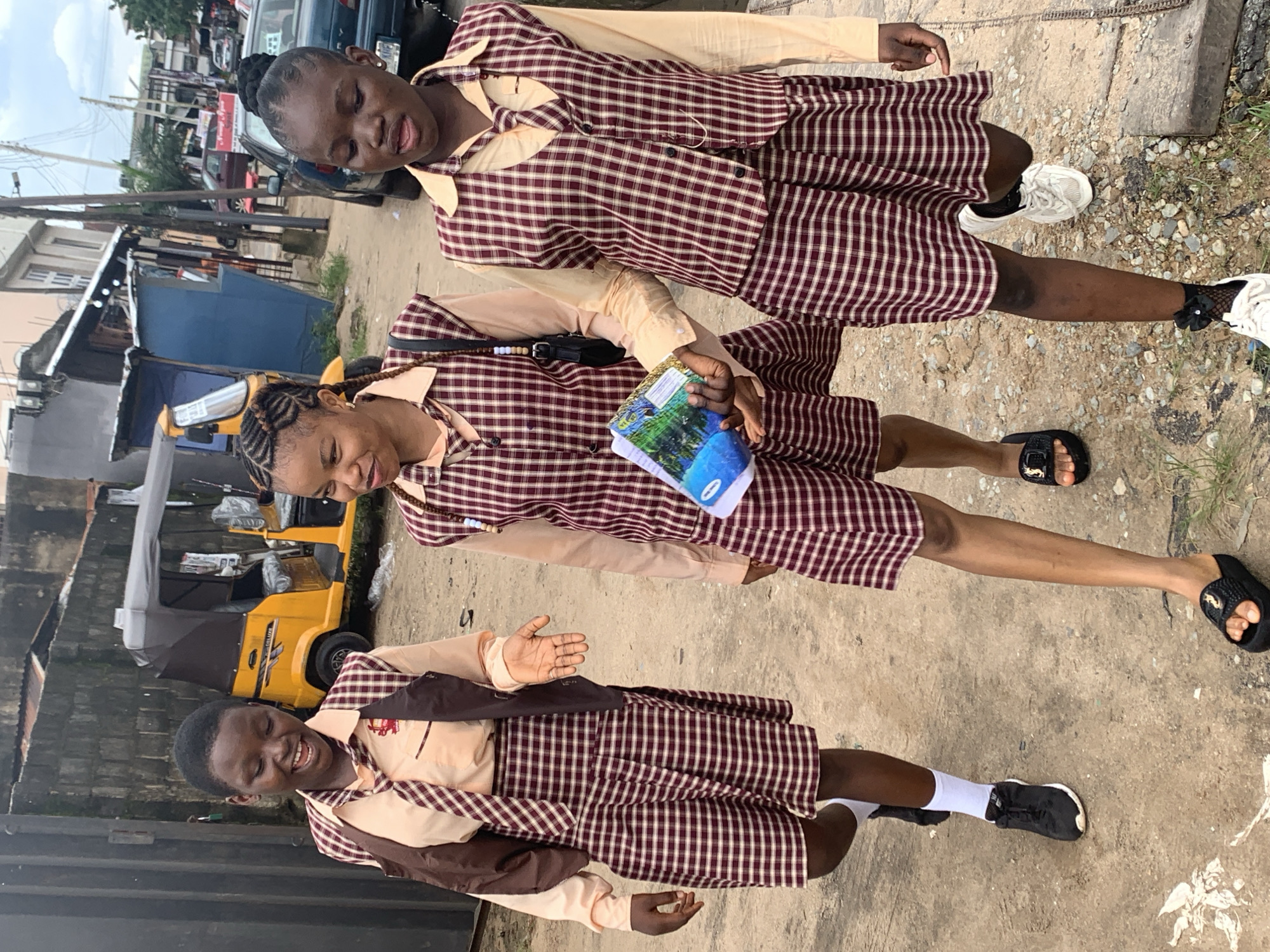 Three school-aged girls in matching maroon and white plaid uniforms, including shirts, ties, vests, and skirts, walk along a dirt path in Umuode, Nigeria. The girl on the left, wearing black sneakers and white socks, laughs as she gestures with her left hand. The girl in the center, carrying a booklet or binder, looks forward with a smile. The girl on the right also looks forward and has beads around her neck, wearing black slides. In the background, a yellow auto-rickshaw is parked near a building with a blue facade. Other buildings and parked vehicles are visible further down the street, suggesting an urban or semi-urban setting. The ground is a mixture of dirt, pebbles, and scattered debris.