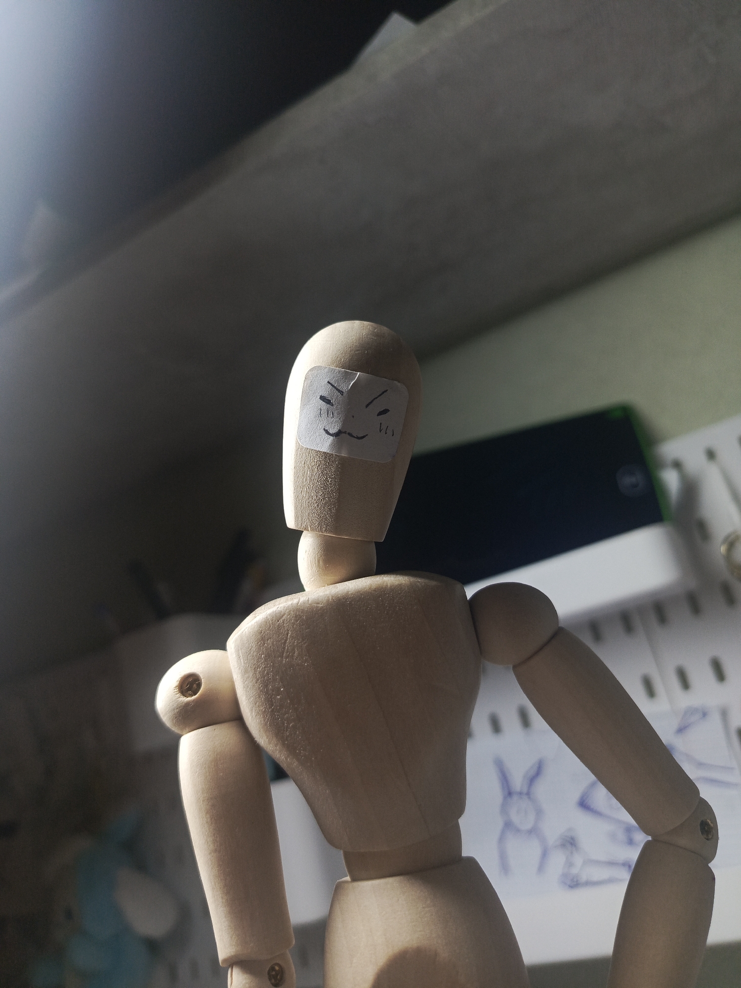 A close-up shot shows a wooden art mannequin wearing a bandage on its face with a drawn expression. The mannequin is positioned slightly off-center, facing the viewer, with the camera angled upwards. The background features a shelf with various items, including a black electronic device and sketches, one of which appears to be a drawing of a rabbit. The environment is dimly lit, creating shadows and highlighting the mannequin. The location is not discernible from the image.