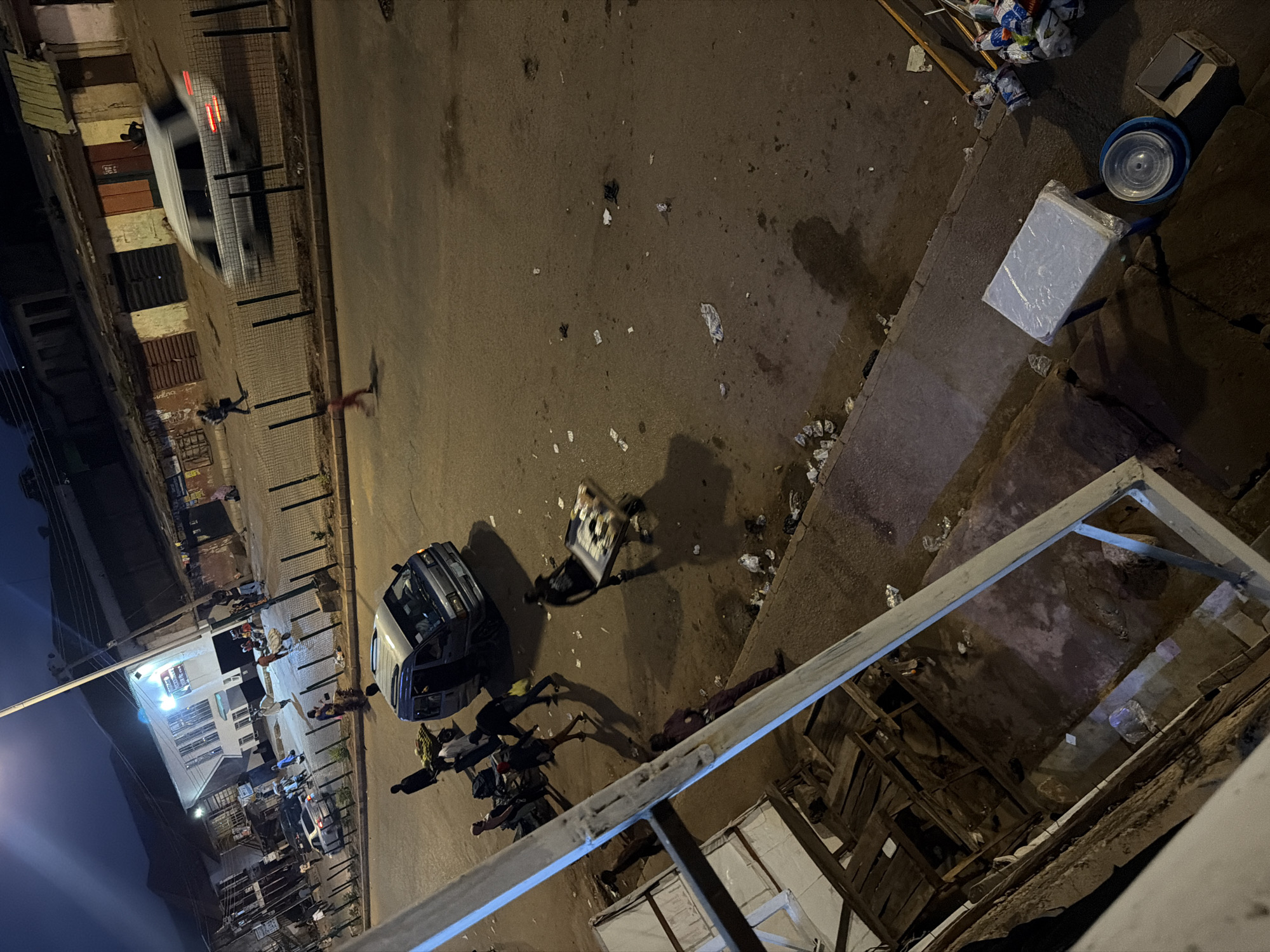 This is a high-angle, medium shot of a street scene at night in Bukuru, Nigeria. The street is paved and appears wet, with scattered debris. On the right side of the street, there is a light-colored van with its doors open, and several people are gathered around it. One person is pushing a cart laden with goods, while others stand or walk nearby. Further down the street, a white car is in motion, and another person is walking. On the left side of the image, a structure with wooden planks is visible, possibly a market stall or temporary shelter. The scene suggests a bustling street market or a busy thoroughfare at night, with commercial activity and pedestrian traffic.