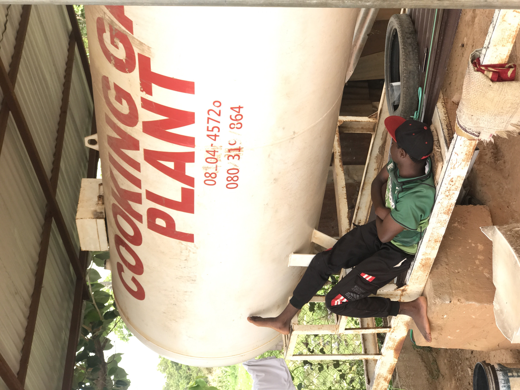 A large, white cooking gas tank with the words "COOKING GAS PLANT" in red lettering, along with the phone numbers "08104-45720" and "080-310/864", dominates the scene. A young Black man, wearing a green shirt, black pants with red and white stripes, and a red and black baseball cap, is sitting on a rusty, makeshift metal structure attached to the tank. He has his arms crossed and appears to be looking upwards. A spare tire and a patterned cloth are visible on the metal structure behind him. The scene takes place outdoors, as green foliage and trees are visible in the background. Metal roofing can also be seen. The location is most likely Abuja, Nigeria, due to the phone number format.