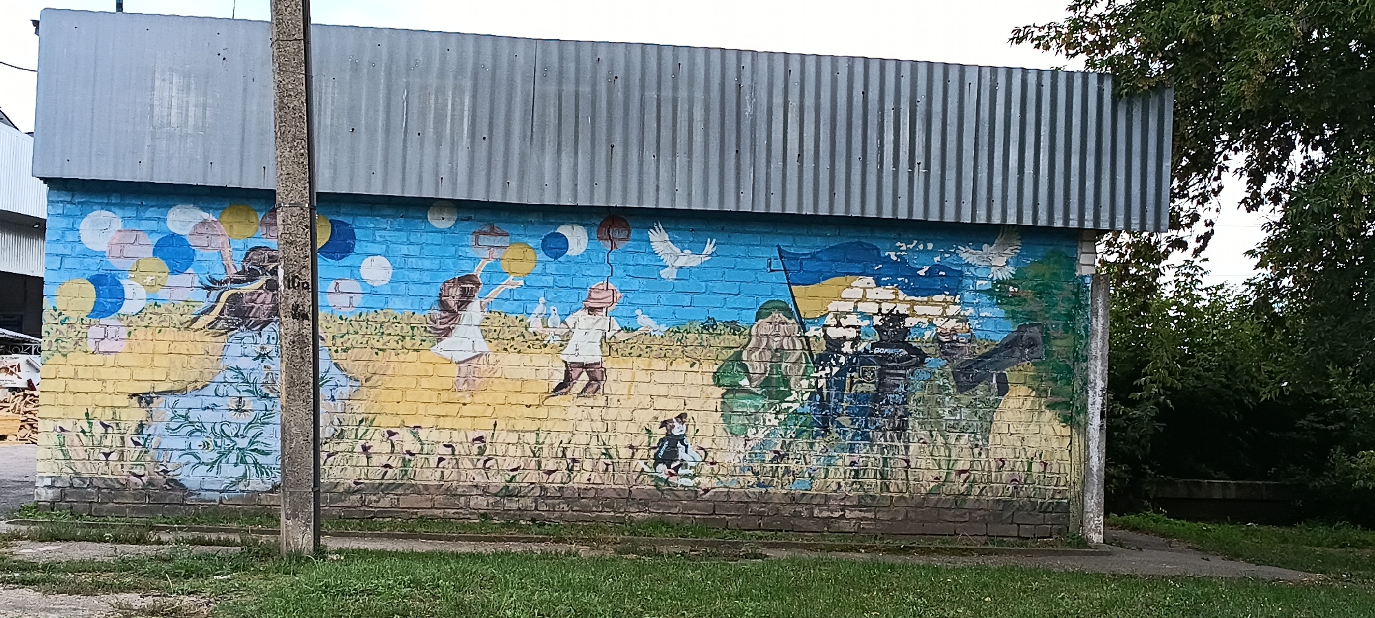 This image shows a mural painted on the side of a brick building in the city of Kamianka, Ukraine. The mural depicts a scene with several elements. On the left, a figure in a blue and white dress is reaching up towards a cluster of colorful balloons. To the right of this figure, two children are shown, one in a white dress holding a yellow balloon and the other in a hat and brown clothing, with several small birds nearby. Further to the right, a Ukrainian flag is depicted. Below the flag, several figures in military-style uniforms are present, with one figure wearing a helmet and the word "ПОЛІЦІЯ" (Police) visible on their uniform. There are also two white doves flying in the sky portion of the mural. The building itself has a corrugated metal roof. The mural is painted over a brick wall, and some areas of the paint are chipped or faded. In the foreground, there is green grass and a concrete path. A concrete utility pole stands in front of the mural, partially obscuring it. Lush green trees and foliage are visible behind the building on the right side of the image.