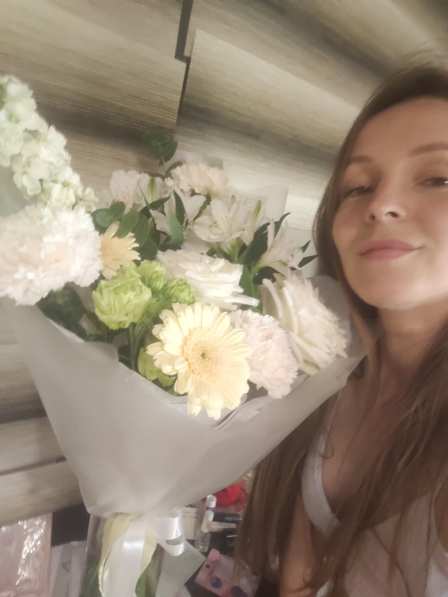 A person with long brown hair holds a bouquet of white and yellow flowers wrapped in white paper with a white ribbon. The bouquet includes carnations, gerbera daisies, roses, and other white flowers. The background is a wall with a striped pattern. The location is Chaikovsky, Russia.