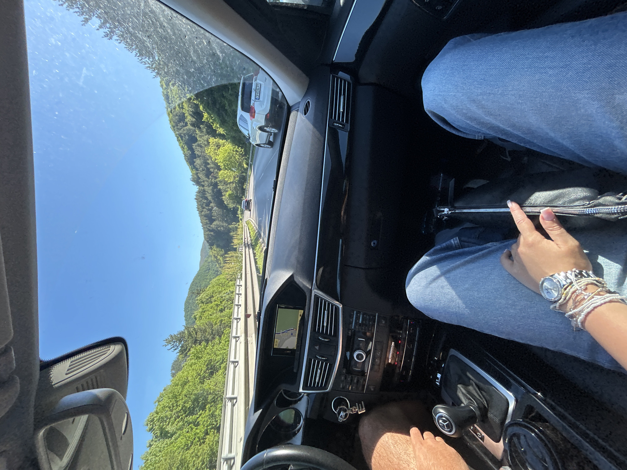 The image shows the interior of a car driving on a highway with green foliage lining the road. The scene is viewed from within the vehicle, and a dashboard with buttons and a navigation screen is visible. In the passenger seat, an arm and leg clad in blue denim are seen. The location is Churek, Bulgaria, inferred from context. There are no discernible actions or interactions between people. The general environment is sunny and bright, with a clear sky overhead. A vehicle can be seen driving in front of the car.