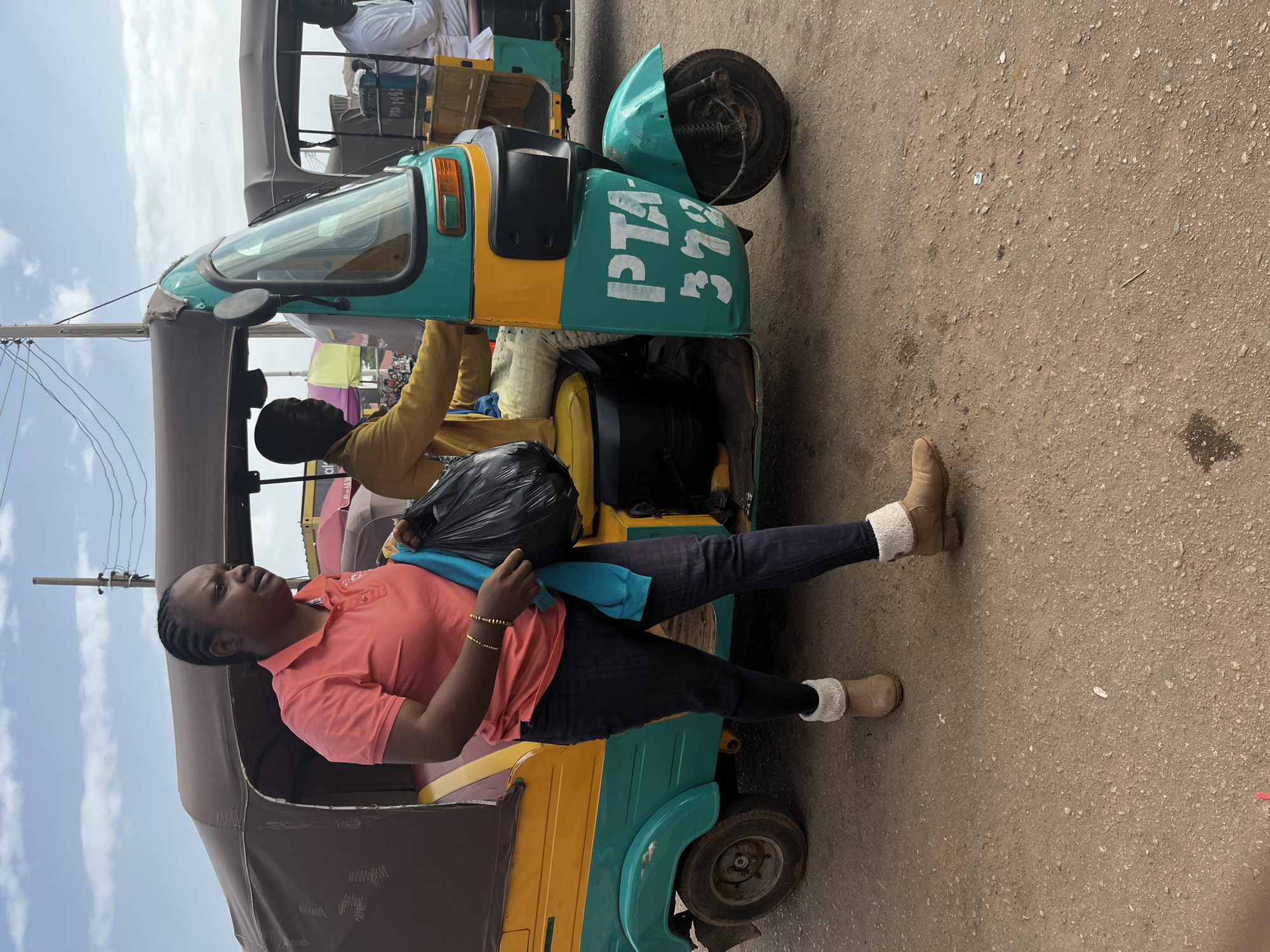 A woman in a pink polo shirt and dark jeans walks past a teal and yellow auto-rickshaw in Gura Top, Nigeria. She is carrying a black plastic bag and a blue garment over her left arm. The auto-rickshaw has "PTA 378" painted on its side. A man in a yellow shirt is visible in the driver's seat of the auto-rickshaw. The ground is paved with gravelly dirt. The sky is cloudy.