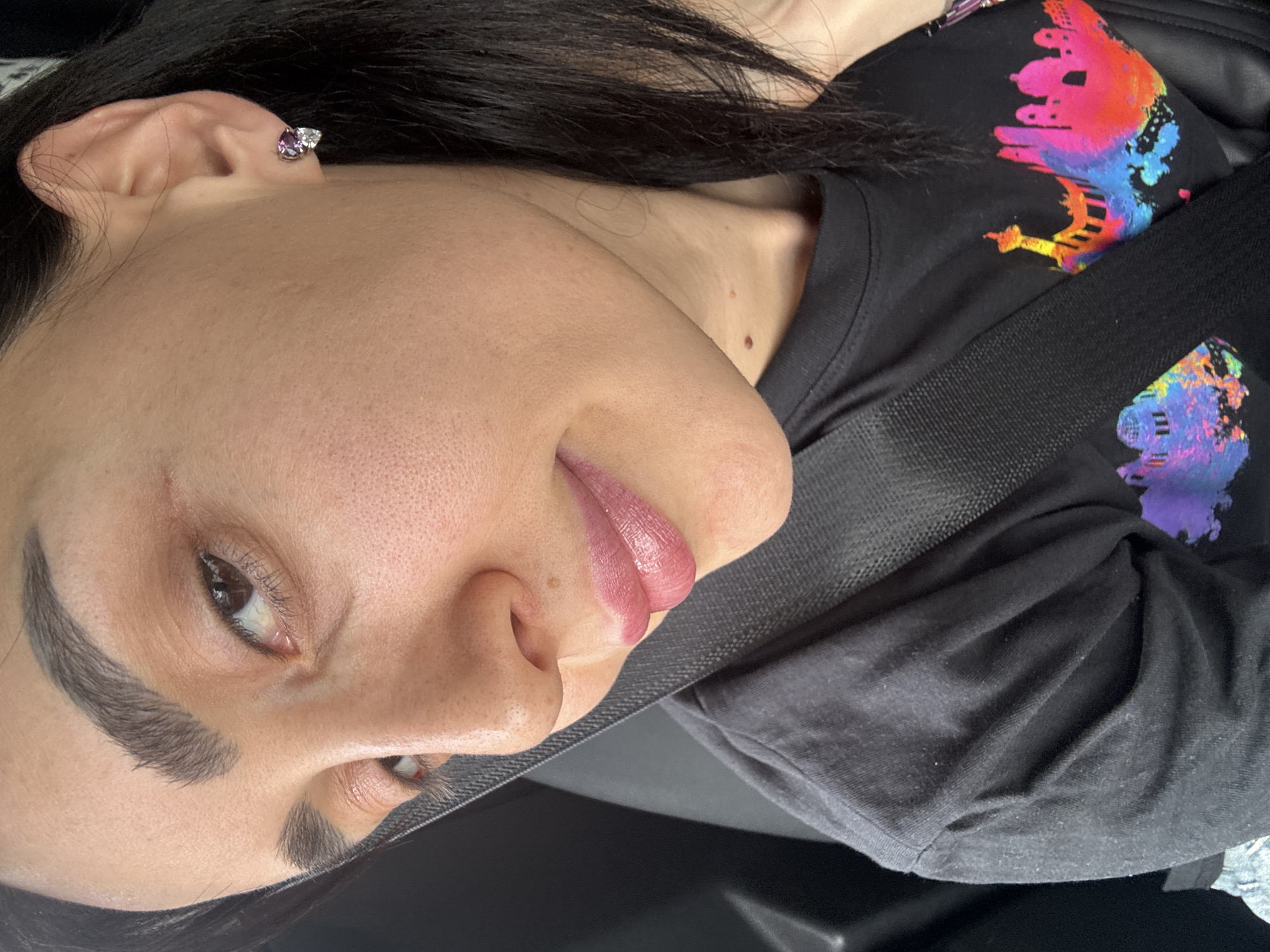A close-up, slightly angled shot features a person with fair skin, dark hair, and dark eyebrows. They are wearing a black t-shirt with a colorful design of Tbilisi landmarks on it. A seatbelt is slung across their body. The person has an earring in their left ear. The location is likely Tbilisi, Georgia due to the shirt's design.