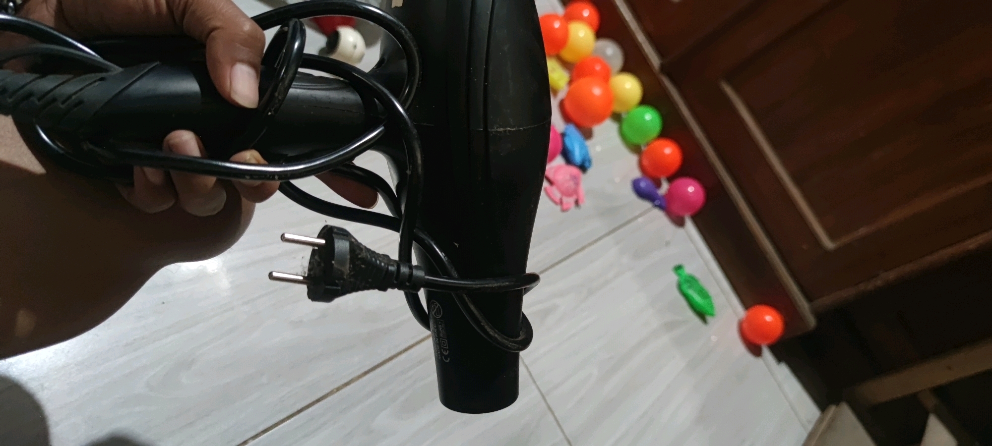 A hand holds a black hair dryer with its power cord wrapped around the barrel and a dirty plug resting against the body of the appliance. The hair dryer is positioned on a light-colored tiled floor. In the background, scattered on the floor, are several brightly colored plastic balls, including red, yellow, green, blue, and pink. A portion of a wooden cabinet or door frame is visible on the right side of the image. The scene appears to be indoors.