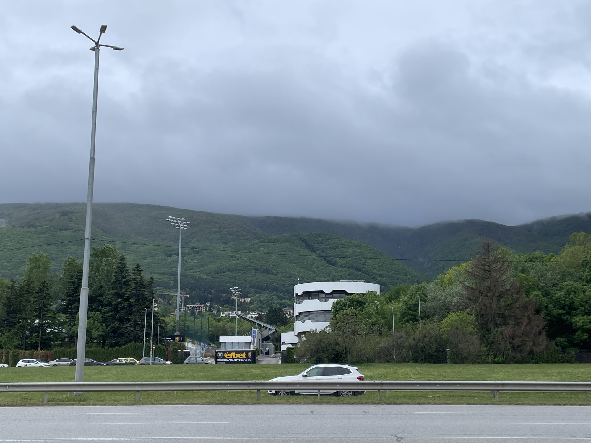 The image depicts a street scene in Sofia, Bulgaria. A grey roadway extends across the bottom of the frame, bordered by a metal guardrail and a grassy median. Beyond the median, several cars are parked near a soccer stadium branded with "efbet" signage. A modern, white, cylindrical building with horizontal banding stands amidst the greenery. In the background, a mountain range is shrouded in low-hanging, grey clouds. The sky is overcast, creating a diffused light. A tall, slender lamppost stands on the left side of the frame. The scene is predominantly green and grey.