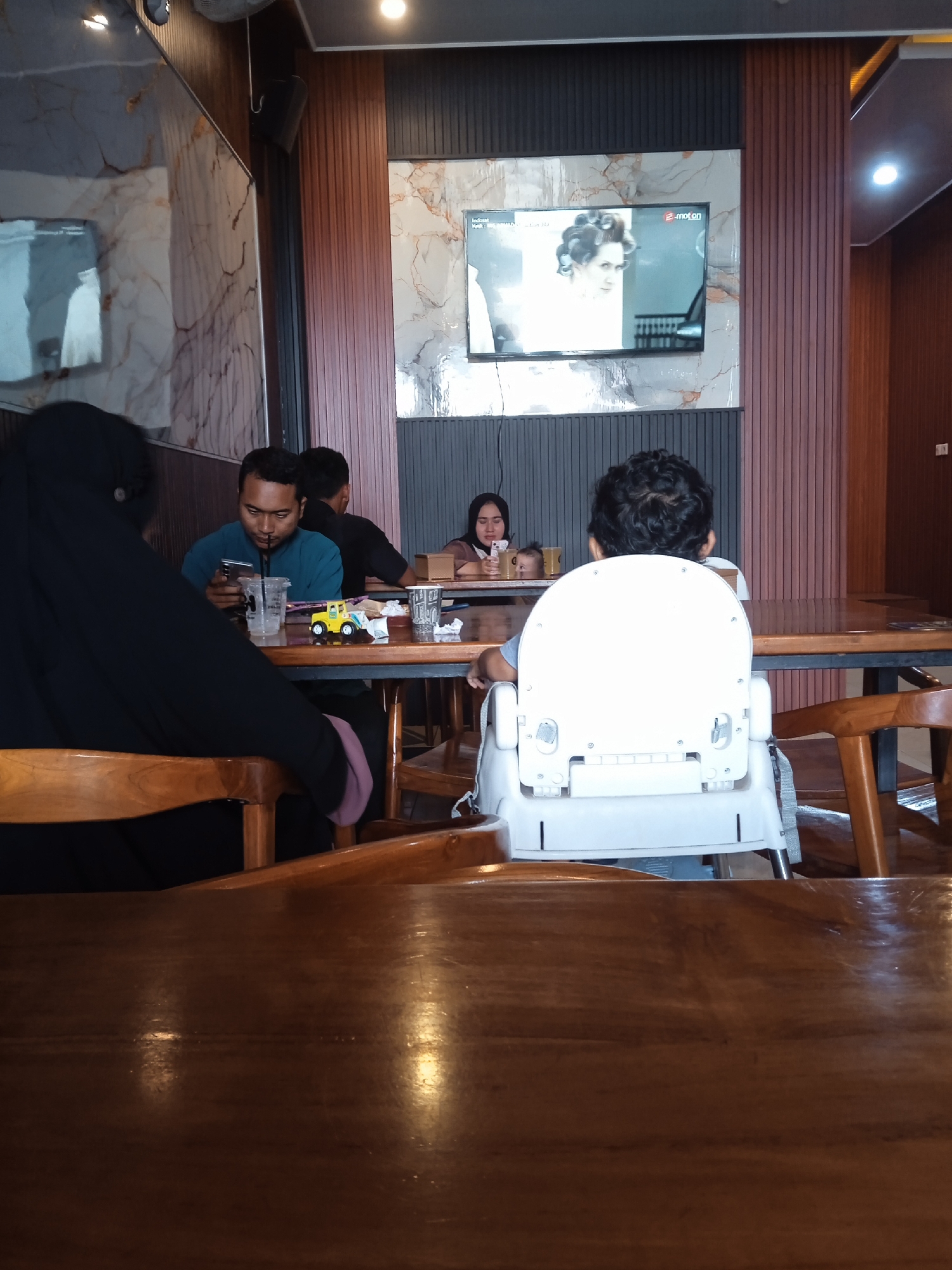The photograph, taken indoors in Pandean, Indonesia, captures a family gathering in a café setting. In the foreground, a glossy wooden table dominates the view. In the midground, a man in a blue shirt is focused on a smartphone, next to a clear plastic cup with a straw, a small toy car, and other items. To the left, a woman wearing a black niqab is seated. In front of the man, a child sits in a white highchair. In the background, a woman is holding a baby. A television mounted on the wall above displays a black and white image of a woman. The café's decor includes patterned wall panels in shades of grey and brown. The overall atmosphere suggests a casual family outing.