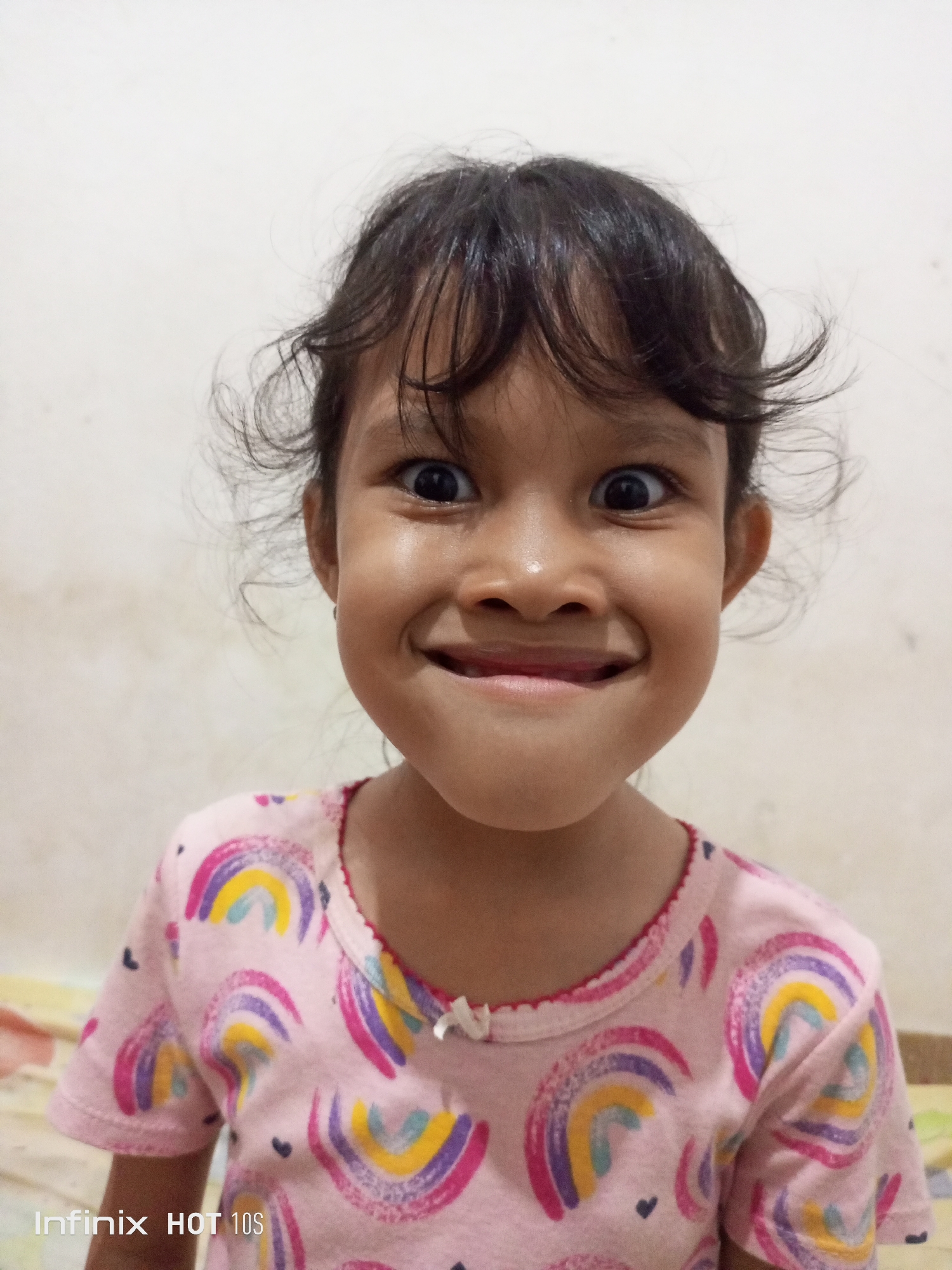 A young girl is shown from the shoulders up, looking directly at the camera with a wide, playful smile. Her eyes are wide and her hair is slightly messy. She is wearing a pink t-shirt with a pattern of colorful rainbows. The background is a plain, light-colored wall. The bottom left corner of the image shows the text "Infinix HOT 10s" which is likely a watermark from the device used to take the photo. The overall impression is one of joy and innocence.