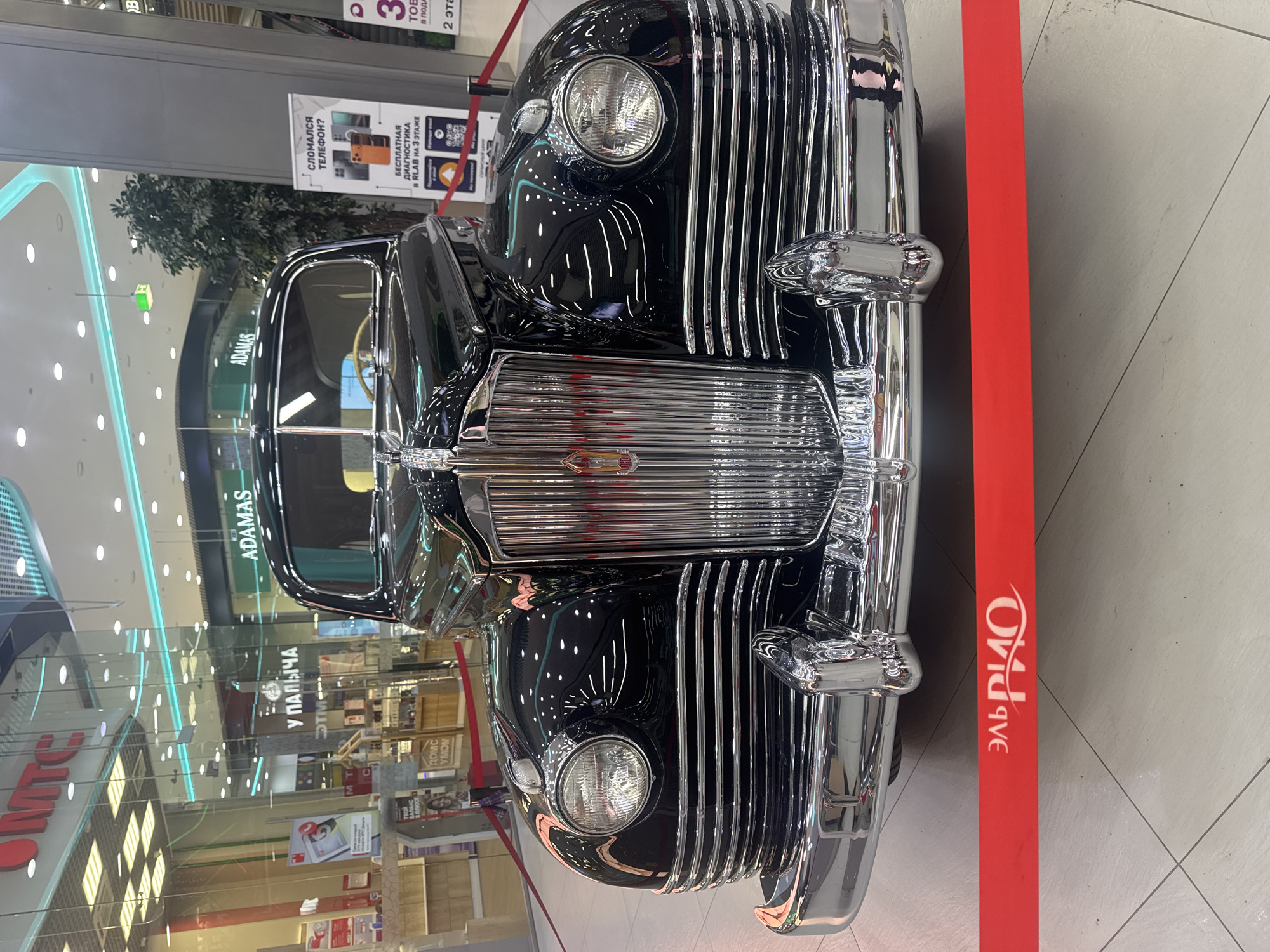 This is a vertical, eye-level medium shot of the front of a classic black car, likely a Soviet-era ZIL or GAZ. The car is highly polished and reflects the surrounding environment, including fluorescent lights and display windows. The scene appears to be indoors, possibly in a shopping mall, given the presence of retail signage and the polished flooring.
The car's front grille is prominent, a large, chrome-plated vertical bar design. Two large circular headlights are visible on either side of the grille, also featuring chrome bezels. The car's hood slopes down towards the grille.
In the background, several signs are visible. One sign, in Russian, reads "СЛОМАЛСЯ ТЕЛЕФОН?" which translates to "Phone broken?". Below this, another sign advertises "БЕСПЛАТНАЯ ДИАГНОСТИКА В RLAB НА 3 ЭТАЖЕ", meaning "Free diagnostics at RLAB on the 3rd floor". Other Cyrillic text on signs includes "ADAMAS" and what appears to be "У ПАЛЫЧА". A red barrier tape with the white Cyrillic text "ЭЛЬВИО" runs vertically along the right side of the frame.
The overall impression is of a car exhibition or a display within a commercial space. The lighting is bright, suggesting it's daytime, and the atmosphere appears clean and modern, contrasting with the vintage vehicle. There are no people clearly visible in the frame.