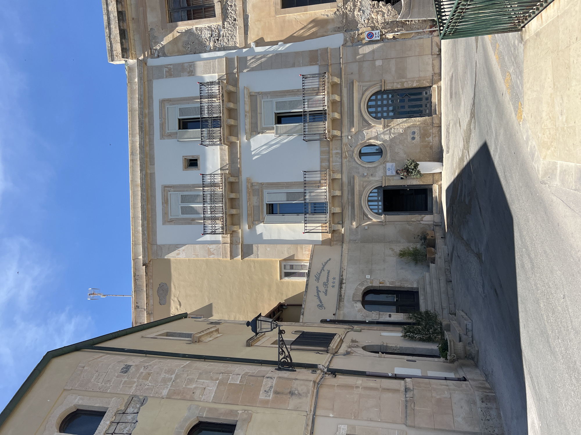 The image depicts a street scene in Syracuse, Italy, featuring the exterior of "Residenza Alberghiera dei Baroni". The architecture is characteristic of the region, with buildings constructed from light-colored stone. In the foreground, a street slopes upwards, casting shadows from the buildings onto its surface.
The main building in focus is a multi-story structure with a light-colored facade. It has several windows with dark shutters and small balconies with wrought-iron railings. Above one of the entrances, an inscription reads "Residenza Alberghiera dei Baroni" with three stars below it, indicating it is a hotel or lodging. The entrance itself is reached by a short set of stairs. Potted plants and a decorative urn are placed near the entrance.
To the left of this building, another structure with a yellowish facade is visible, featuring arched windows and a wrought-iron lantern mounted on a decorative bracket. Further to the left, a glimpse of another building with stone cladding can be seen.
The sky above is clear and blue, with a few distant birds visible. The overall impression is of a sunny day in a historic urban setting.