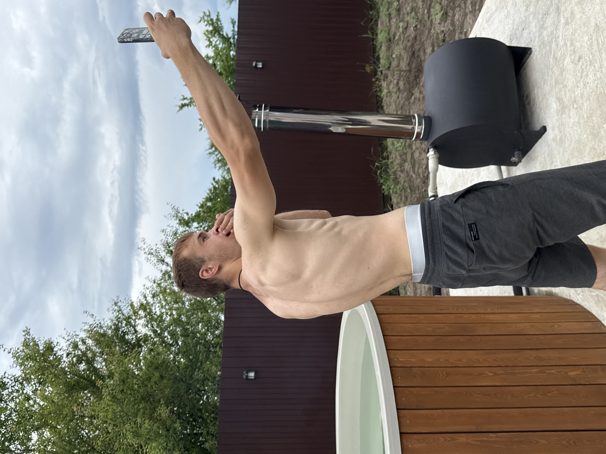 A shirtless young Caucasian man with short brown hair is taking a selfie outdoors. He is wearing dark gray sweatpants with a light gray waistband. He is standing to the right of a wooden hot tub filled with greenish water and to the left of a black metal wood-fired oven with a stainless steel chimney. Behind him is a dark brown wooden fence with a light fixture on it, and beyond that, trees and a cloudy sky. The man's left arm is extended, holding a mobile phone, and his right hand is near his mouth. The scene appears to be in the city of Starokorsunskaya, Russia, possibly a backyard or a recreational area.