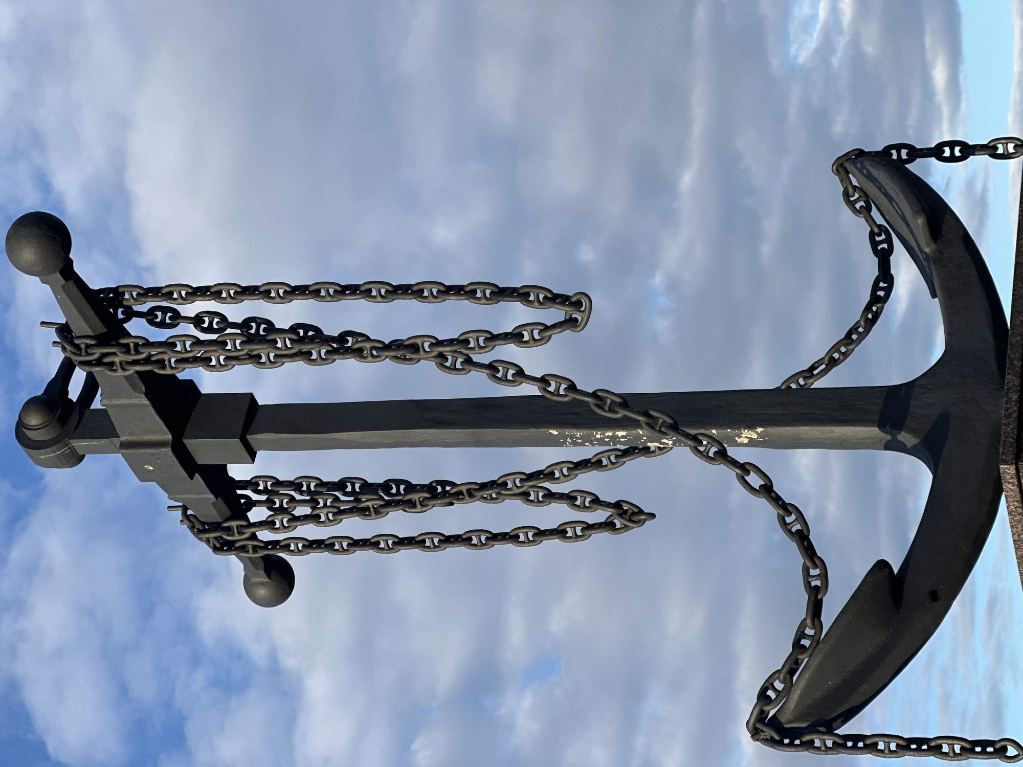 An iron anchor with chains hangs against a cloudy sky. The anchor is black, and the chains are dark grey. The anchor's stock, with spherical ends, runs horizontally across the frame. The anchor's shank is vertical. The sky is mostly light blue with some white clouds.
The anchor is a monument in Votkinsk, Russia.