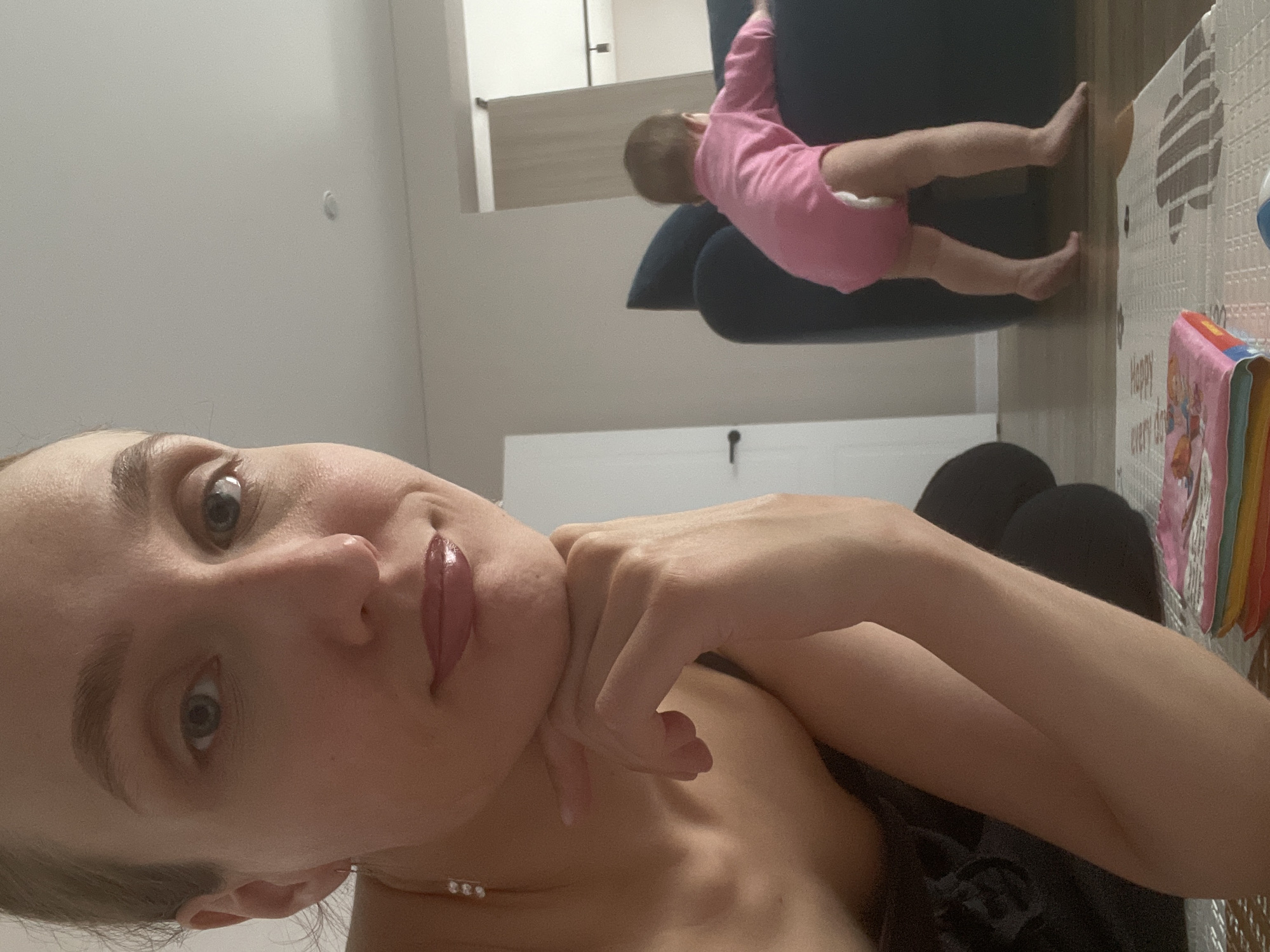 The image is a tilted, low-angle indoor selfie featuring a woman in the foreground and a baby in the background, set in Sepych, Russia.
In the foreground, an adult woman with light skin, light-colored eyes, and glossy, reddish lips looks directly at the camera with a calm, slightly smiling expression. Her light hair is mostly out of frame. She wears a dark, thin-strapped top and a delicate necklace with three small, sparkling elements. Her right hand rests under her chin, with fingers curved around her jawline.
In the mid-ground, a baby or toddler is seen from behind, wearing a pink long-sleeved onesie and a white diaper. The baby is on a dark blue sofa or large cushion, appearing to be climbing or standing, with its bare legs and feet visible.
The background shows light-colored walls. To the right of the baby, a patterned foam play mat is on the floor. A colorful children's book or toy with "Happy every d" (likely "Happy everyday") printed on it is next to the mat. A small, round, white object is visible on the wall, and reflections of a mirror or glass surface can be seen further back. A small black handle or knob is present on a white surface in the lower background.
The image captures a casual, domestic moment, likely a parent's selfie with their child playing nearby. The lighting suggests an indoor setting, possibly during the day or evening.