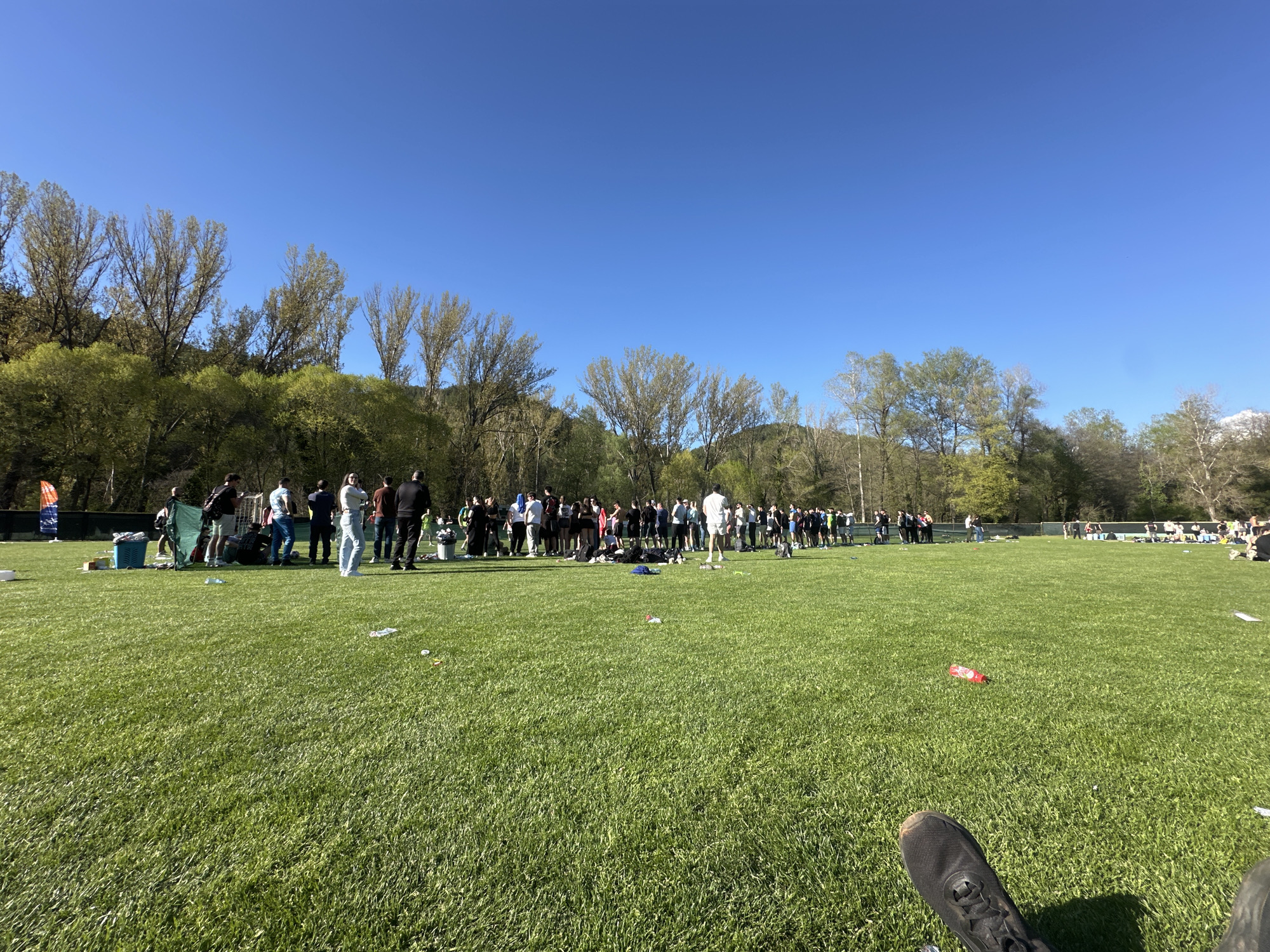 The image shows a large group of people gathered on a grassy field under a clear blue sky, likely in Blagoevgrad, Bulgaria. The crowd appears to be a mix of individuals, possibly participants and spectators of an event, as they are standing in a somewhat organized line or cluster. The surrounding environment includes trees and some low-lying hills in the background, suggesting a park or recreational area. There is some scattered trash on the grass. In the foreground, a foot with a black shoe is visible, indicating the perspective of the photographer.