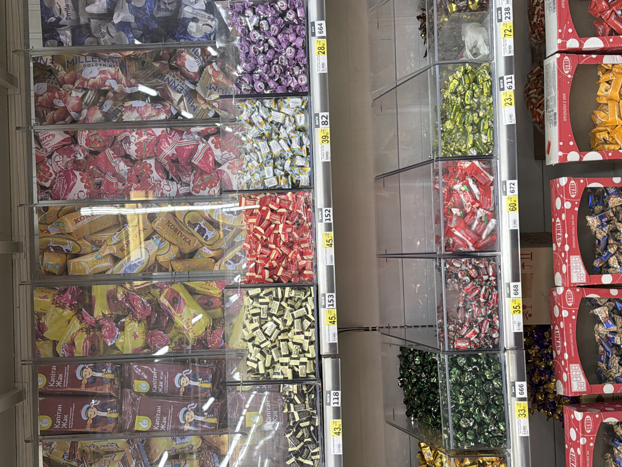 The image shows a retail display of bulk candies in plastic bins at a supermarket in Sokilnyky, Ukraine. The candies are of various colors and wrappers, including red, yellow, purple, and green. Some of the wrappers have text that is difficult to read but appears to be in Ukrainian. Below the bins are price tags with currency and numbers, indicating prices per unit weight. The environment appears to be an indoor market or grocery store with overhead lighting and metal shelving. There are no people visible in the image. The overall scene is one of a commercial setting focused on the sale of confectionery.