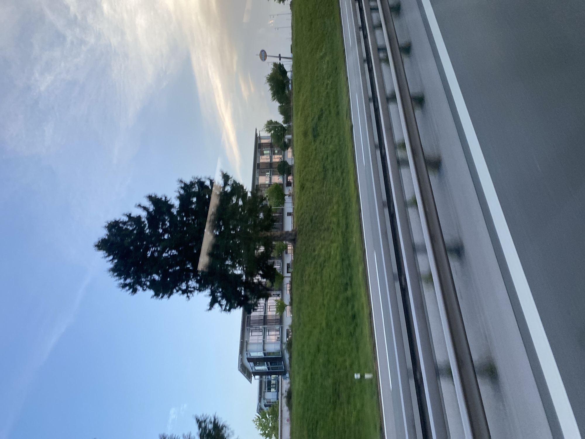 The image shows a modern office building with a glass facade, situated on a grassy hill. A large evergreen tree stands in front of the building, partially obscuring it. The building appears to be multi-story and has a flat roof. The scene is viewed from the perspective of a vehicle traveling on a highway, with guardrails and road markings visible in the foreground. The sky is partly cloudy, with a mix of blue sky and white clouds, suggesting it is either early morning or late afternoon. A sign with the "Fossil" logo is visible to the right of the building. The location is identified as Grabenstätt, Germany.