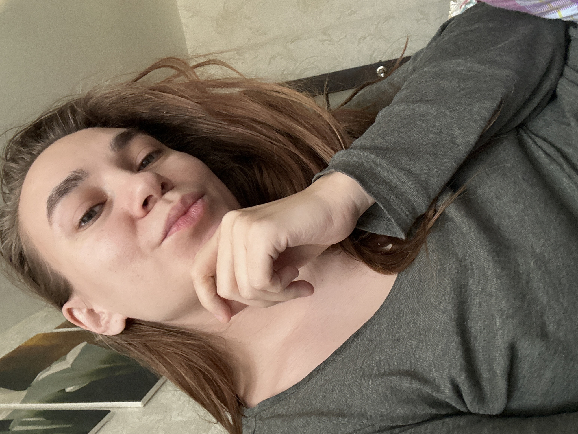 A woman with light skin and brown hair is angled towards the camera. She is wearing a gray long-sleeved shirt. Her right hand is near her face, and she appears to be lying down. In the background, part of a painting or framed artwork is visible on the wall, which is also covered in light-colored patterned wallpaper. The location in the city of Uva, Russia is not discernible from the provided image.