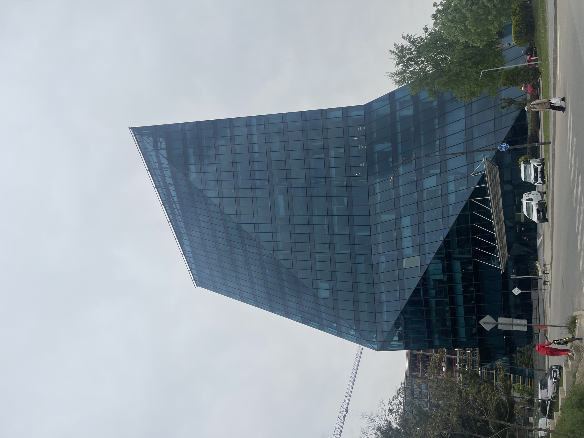 The image shows a tall, modern office building made of glass in Sofia, Bulgaria. The building has an angular, geometric design and is primarily blue-tinted. The sky is overcast. A street runs along the side of the building, with trees and some pedestrians visible. Several parked vehicles are present. There is also a crane visible near the bottom of the building, suggesting construction work nearby. A street sign is adjacent to the road. A second, smaller building is visible to the lower right of the building in question.