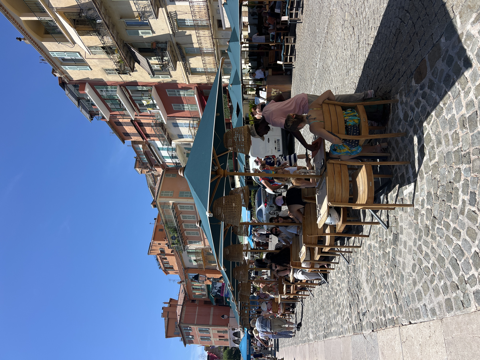 An outdoor seating area of a restaurant is depicted in Villefranche-sur-Mer, France, with patrons seated at tables under large green umbrellas. The tables are made of wood with metal legs, and wooden chairs surround them. The ground is paved with cobblestones. In the background, a row of colorful buildings with multiple stories and balconies lines the street. A white van is parked to the left of the frame. People are scattered throughout the scene, some dining, others walking. The sky is clear and blue. The overall impression is a sunny day in a European coastal town.