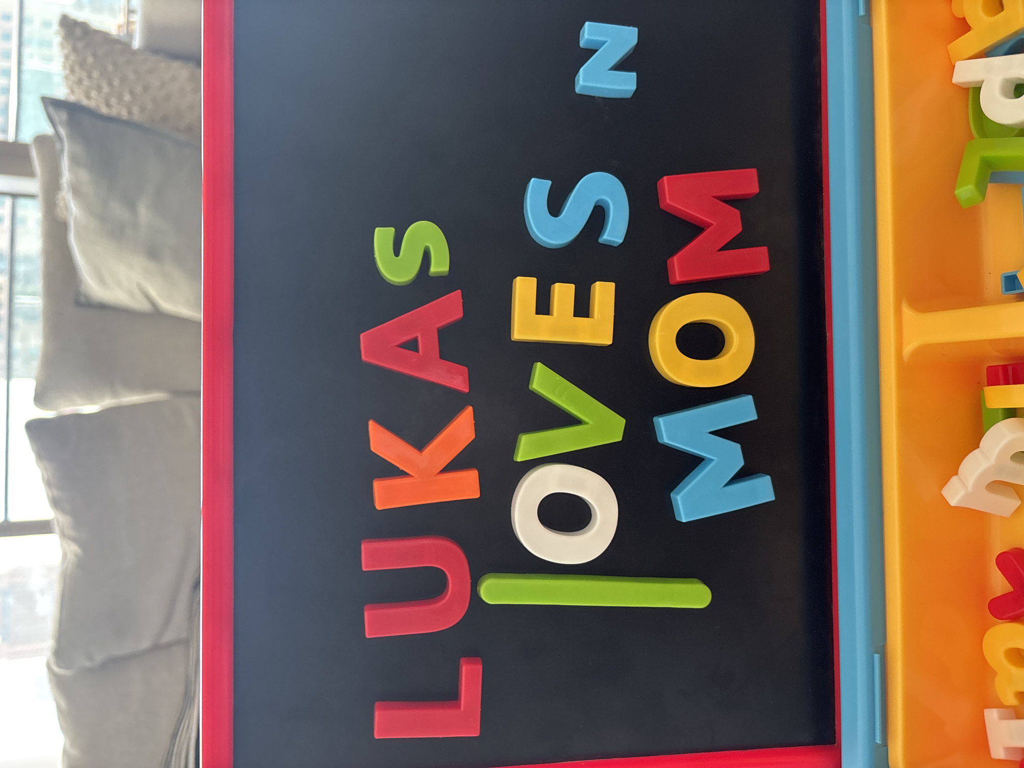 The image displays a close-up view of a magnetic chalkboard with colorful plastic letters arranged to spell "LUKAS LOVES MOM." The letters are red, orange, green, white, yellow, and blue. The chalkboard is framed by red and blue plastic. Below the chalkboard, a section of a toy organizer with orange and blue compartments is visible, containing additional plastic letters. In the blurred background, a couch with light-colored pillows can be seen, along with a window showing daylight. The scene appears to be indoors, possibly a living room or play area. There are no people present in the image. The specific location in Dubai, United Arab Emirates, cannot be determined from the visual information provided.