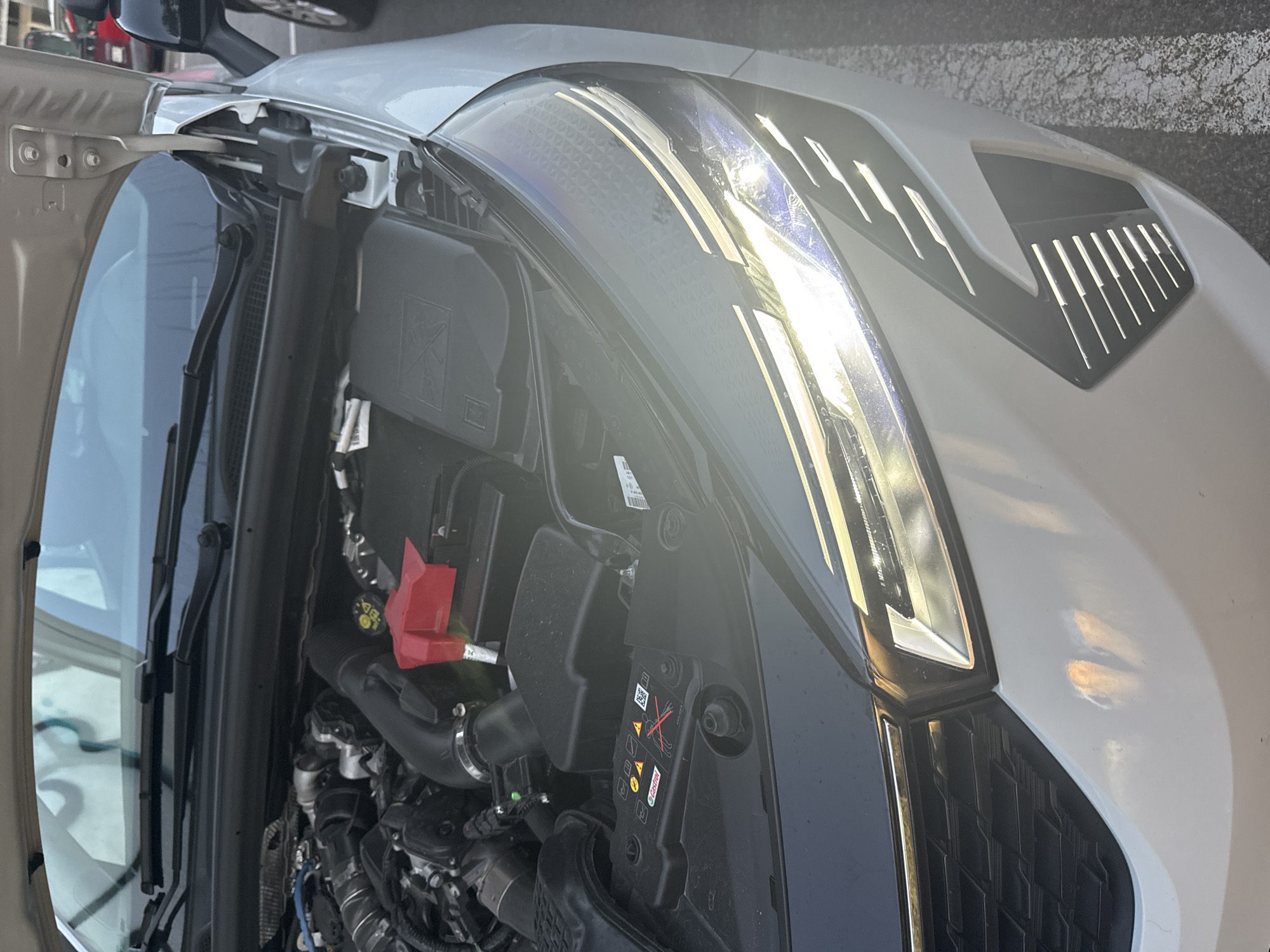 The image shows the front of a white car with its hood open, revealing the engine compartment. The car is parked on a paved surface, possibly a parking lot, with white lines indicating parking spaces. In the background, parts of buildings and other vehicles are visible, suggesting an urban or suburban setting. The car's headlights are on and illuminated, casting a bright light on the front of the vehicle and the surrounding area. The engine bay shows various components, including the battery, air intake system, and wiring. The overall scene appears to be a typical street view in a city.