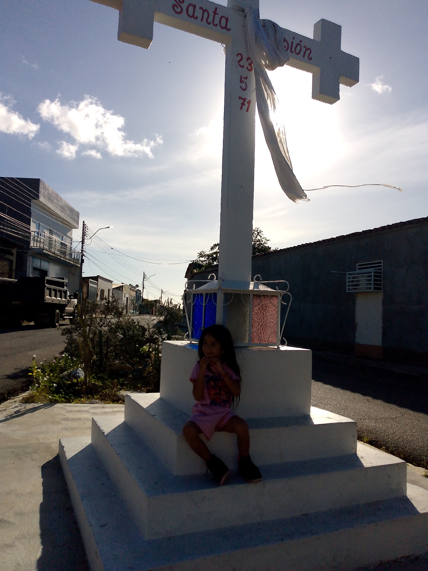 The image depicts an outdoor scene in Juan Griego, Venezuela, characterized by strong backlighting from a low sun. A prominent white concrete cross monument stands centrally on a multi-tiered, stepped base. Red lettering on the cross's horizontal beam reads "Santa" on the left arm and "sión" on the right arm. The vertical shaft displays the red numbers "23," "5," and "71" stacked vertically. A tattered white fabric or cloth piece is draped over the top of the cross, fluttering downwards. Four decorative structures resembling lanterns with blue and pink/red colored glass are affixed to the middle section of the cross's base.
A young girl with dark hair, wearing a pink short-sleeved garment and dark shoes, is seated on the lower steps of the cross's base, positioned to the right of the vertical shaft. Her face is largely in shadow due to the backlighting, and her right hand is held near her mouth or chin.
To the left of the cross, an asphalt street curves into the distance. A dark-colored dump truck is parked or moving on this street. Behind the truck, a multi-story building with a dark lower facade and a white upper section featuring balconies is visible. Numerous overhead power lines connect to utility poles along the street. Further down, additional single-story buildings line the road. Sparse, dry vegetation and some green plants are present along the roadside.
To the right of the cross, another paved area or street runs alongside a long, dark gray wall topped with coping tiles. A small white structure resembling a door or gate with a grated window is set into this wall. The sky is partly cloudy and intensely bright directly behind the cross, creating a halo effect. Long, distinct shadows are cast by the subjects and structures, indicating a low sun angle.
